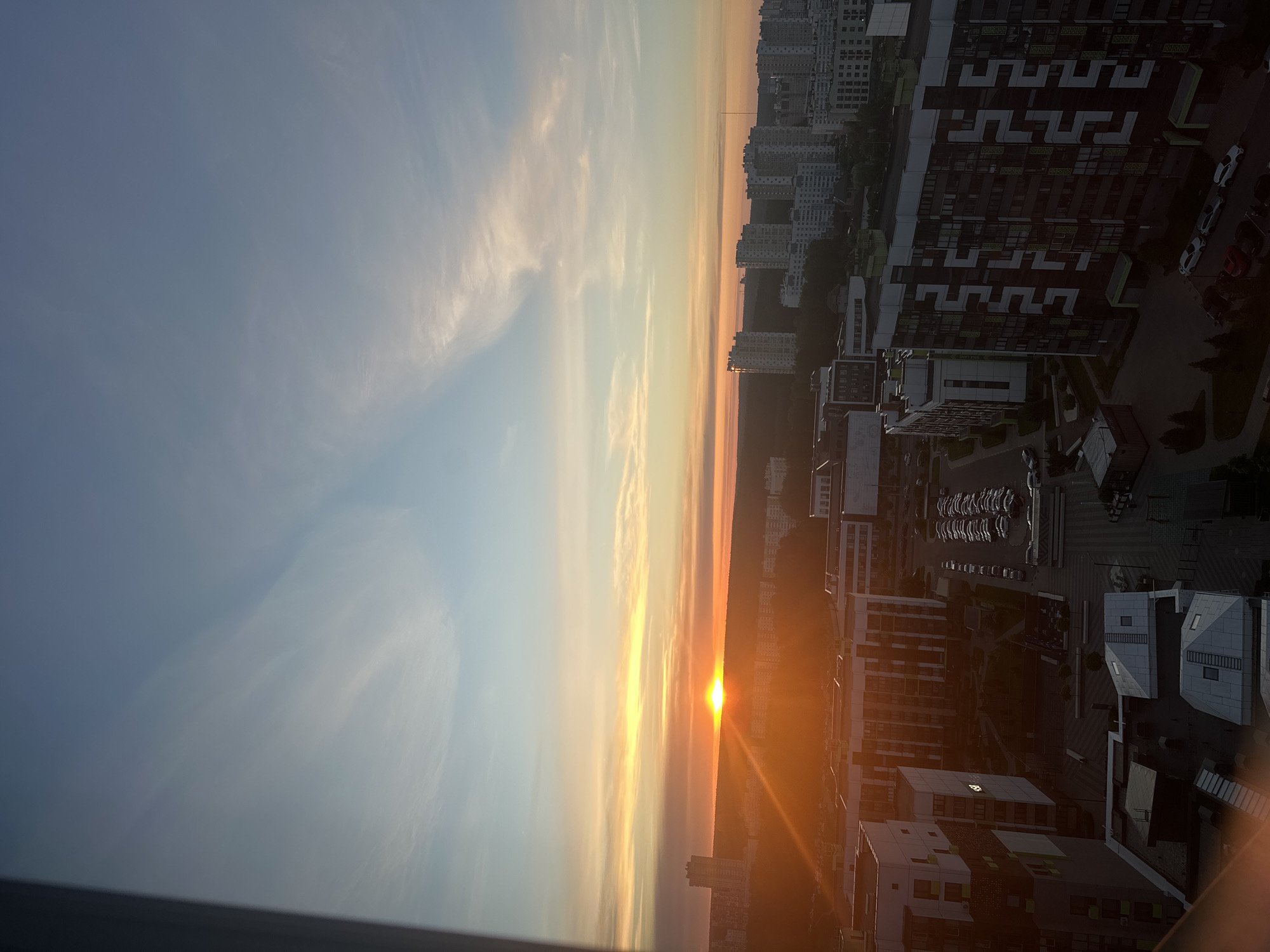 The image depicts a cityscape at sunset in Бараўлянскі сельскі Савет, Belarus. Tall apartment buildings stand on the right side of the frame, their geometric facades casting long shadows. The sun, a bright orange disc, is partially obscured by the horizon. The sky transitions from blue on the left to a hazy orange near the horizon. Clouds are visible in the sky.
