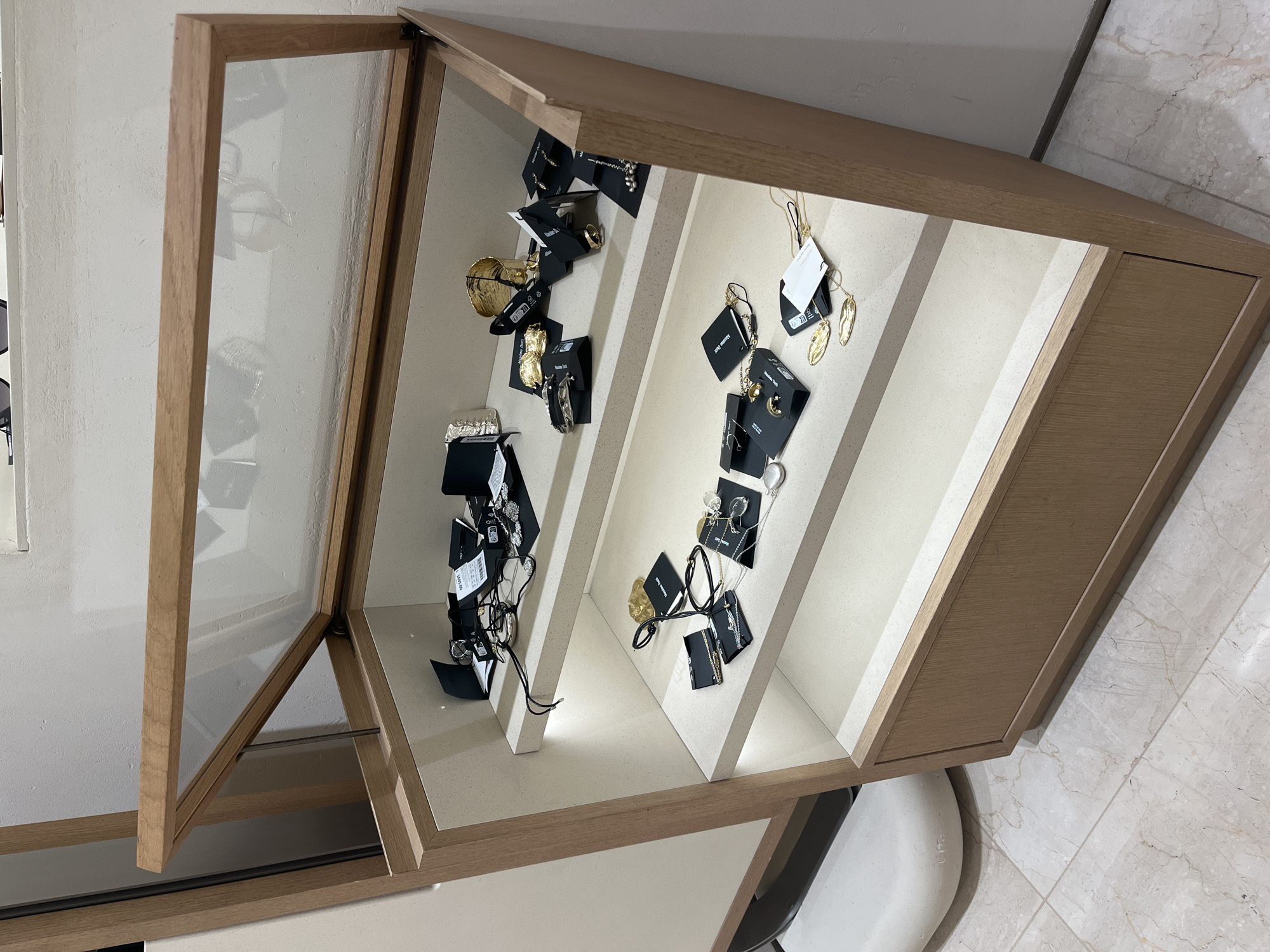 A display case, constructed from light-colored wood, showcases an array of jewelry items attached to black tags. The case is positioned on a surface, with a white object visible beneath it. The background includes a white wall, and the floor is covered with beige tiles. Given the style and merchandise, the location may be a retail space in Puebla, Mexico.