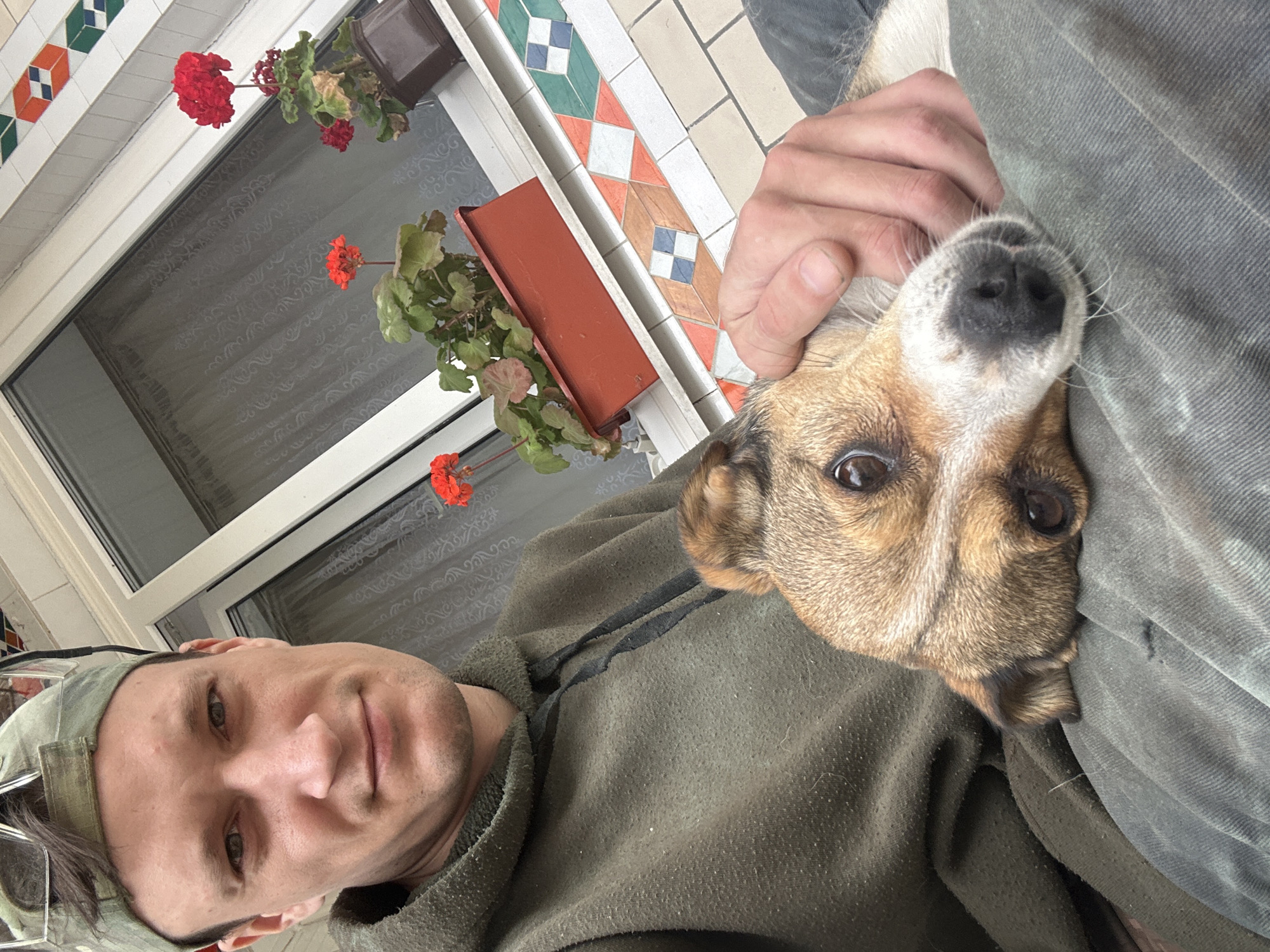 A man wearing a green cap and a green hoodie is holding a dog. The dog is brown and white and is resting its head on the man's lap. The man is looking at the camera and smiling slightly. Behind them, there is a window with white curtains and some red flowers in pots. The scene appears to be outdoors, possibly on a balcony or patio, with a tiled border visible below the window. The overall impression is casual and relaxed. The location is identified as Makiv, Ukraine.