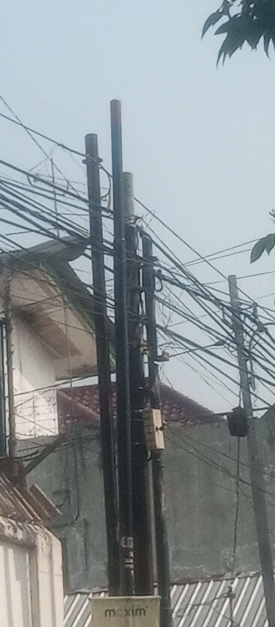 The image shows a utility pole with numerous tangled wires against a hazy sky. In the background, a building with a tiled roof and a light-colored facade is partially visible, along with a concrete wall. The base of the utility pole has a small rectangular box attached to it with the word "maxim" printed on it. The scene appears to be in a developed area, possibly a residential or commercial district in Kediri City, Indonesia, given the presence of utility infrastructure and buildings. The overall atmosphere is somewhat overcast, and the focus is on the complex network of electrical or communication lines.