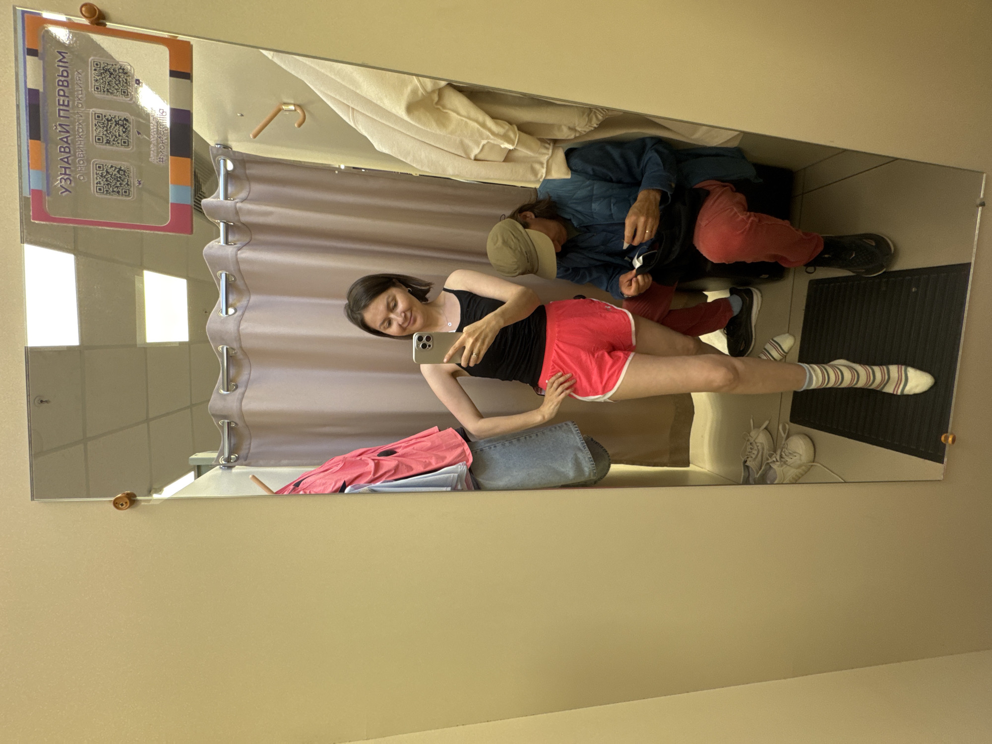 A mirror reflects a woman taking a selfie in what appears to be a dressing room. She is wearing a black tank top and red athletic shorts. An older man wearing a tan hat and blue jacket is seated behind her. A curtain and clothes are visible in the dressing room. A sign with QR codes is attached to the top left corner of the mirror with the word "UZNAVAI PERVYM", which translates to "FIND OUT FIRST". The city of Izhevsk, Russia is not identifiable in this image.