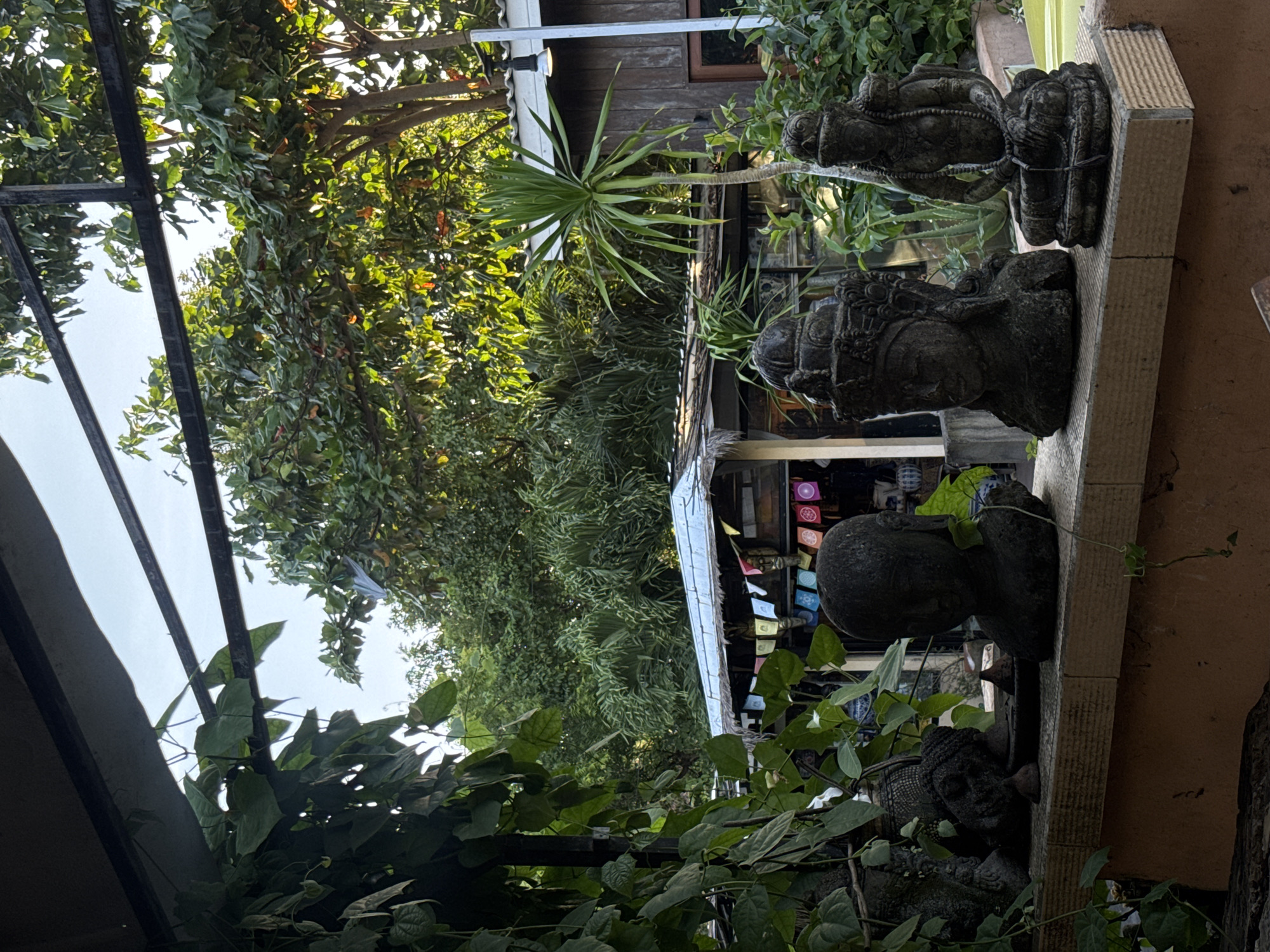 The image, taken in Kuta Selatan, Indonesia, shows a lush outdoor setting with a row of stone statues mounted on a textured, light-colored wall that runs along the right side of the frame. The statues appear to be of deity figures. The area is heavily vegetated with various green plants and trees, some with leafy branches. A structure with a light-colored roof is partially visible in the background, adorned with colorful small flags or decorations. A modern metal frame structure is visible on the left side of the frame, with more vegetation growing up and around it. The overall scene conveys a sense of tranquility and is suggestive of a garden or outdoor space with cultural or religious significance.