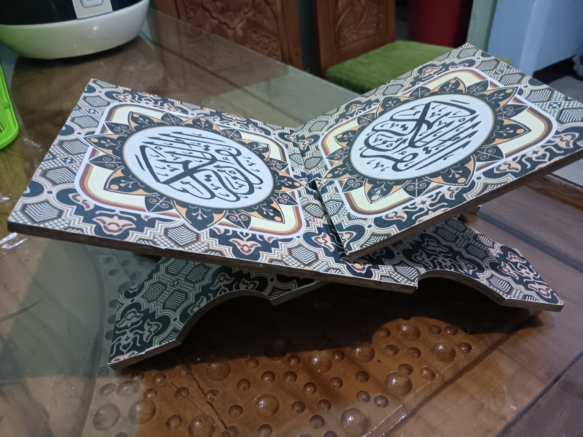 An open book rest on a wooden stand on a glass table in Batursari, Indonesia. The book is decorated with ornate Arabic calligraphy on both pages. The stand has intricate carvings and is also adorned with decorative patterns. In the background, a portion of a white appliance is visible, along with wooden furniture and a green cushion. The table surface features a textured pattern with circular indentations.