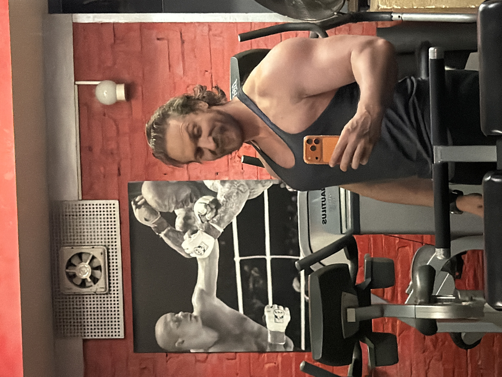 A man is taking a selfie in a gym. He is wearing a tank top and holding an orange smartphone. Behind him is a weight machine and a poster depicting two men boxing. The walls of the gym are red brick. There is a small fan on the left wall and a light fixture above the poster. The photo does not show enough information to determine the location in Saint Petersburg, Russia.
