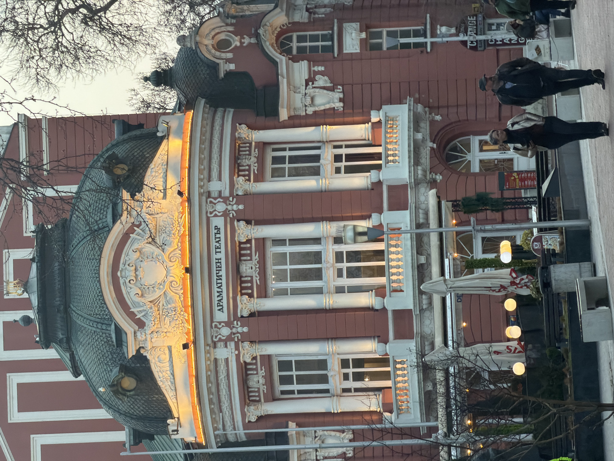 The image shows the facade of the Dramatic Theatre in Varna, Bulgaria. The architecture is classical, with red and white coloring, white columns, and ornate detailing. The building has large windows and a dome-shaped roof with additional ornamentation. A sign in Cyrillic script is visible on the facade.
Two pedestrians are walking on the sidewalk near the building. Street lighting and an outdoor café with umbrellas and globe lights are present. Bare tree branches are visible in the foreground and against the sky, indicating it may be autumn or winter. A "Change" sign is seen in the background, near the pedestrians.