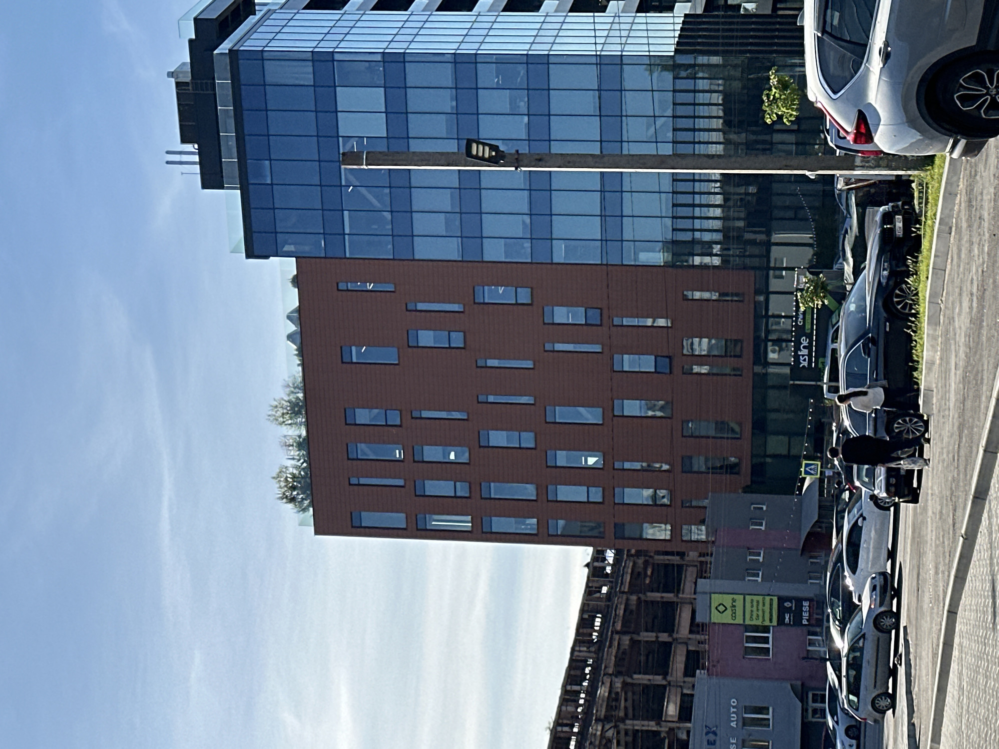 The image captures a city scene with a focus on architecture and urban life. Two modern buildings dominate the background: one with a blue glass facade, the other with a brown facade punctuated by numerous windows. At the base of these buildings, there's a line of parked cars along a paved street. Pedestrians are present, notably a person walking with what appears to be a stroller between the cars. Further down the street, there are more buildings and structures, including what looks like a building under construction with exposed framework. The sky is clear and blue, suggesting daylight. The general location is an urban area with modern architecture and signs of ongoing development.