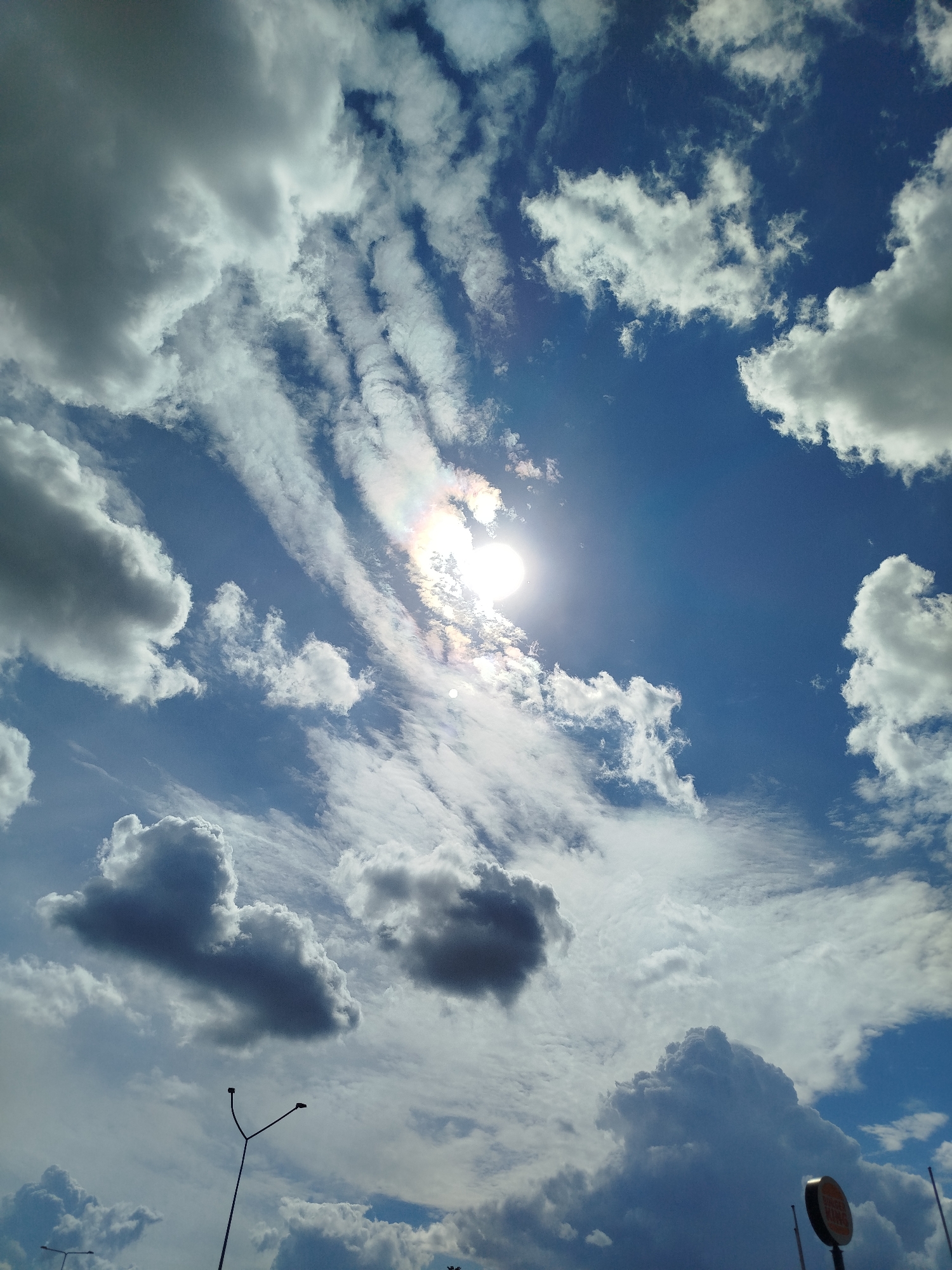The image shows a wide expanse of blue sky filled with various clouds. Some clouds are white and fluffy, while others are darker and more dramatic. The sun is visible, partially obscured by clouds, creating a halo effect with rainbow colors. In the foreground, there are glimpses of what appear to be streetlights or utility poles, suggesting an urban or suburban setting. The overall impression is of a dynamic sky with active cloud formations. This scene is likely located in the city of Ząbki, Poland, a suburban municipality within the Warsaw metropolitan area.