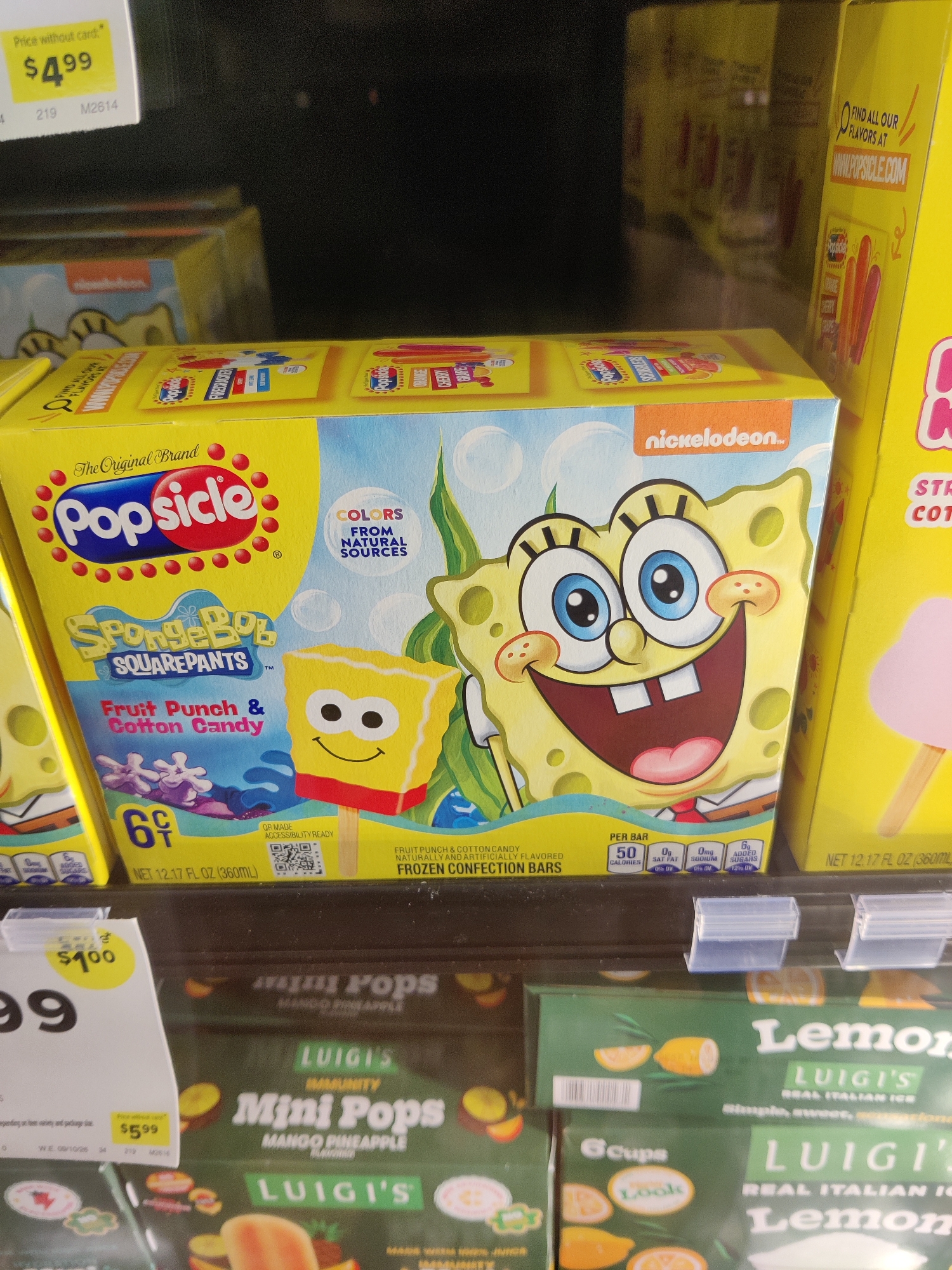 The image shows a close-up of frozen novelty bars in a store freezer. The primary subject is a box of "SpongeBob SquarePants" themed Popsicle bars, featuring Fruit Punch & Cotton Candy flavors. The box is bright yellow with an illustration of SpongeBob SquarePants and his friend Patrick. Nutritional information and branding for Nickelodeon are visible. To the right of the SpongeBob box is another yellow box, likely also frozen novelties, with a pink ice cream bar depicted. Below the SpongeBob box, the top of a green box for "Luigi's Mini Pops" in Mango Pineapple flavor is visible, followed by another green box of "Luigi's Real Italian Ice" in Lemon flavor. The shelves are lined with a white plastic trim. A price tag displaying "$4.99" is visible in the upper left corner of the image, attached to a light yellow label indicating "Price without card." Another price tag showing "$1.00" is on a white label in the lower left. The scene is indoors, in a retail environment, likely a supermarket freezer aisle. There are no people or animals present. The lighting appears to be typical fluorescent store lighting. The specific location of Mechanicsburg, United States of America is not discernible from the visual information in the image itself.