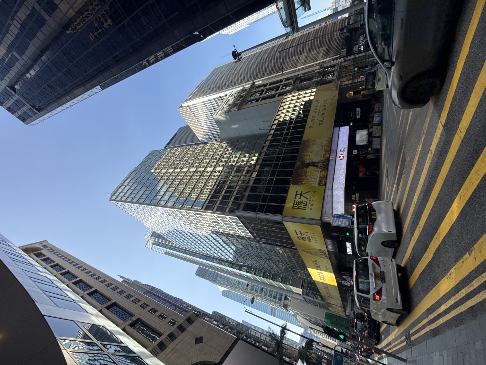 In a wide-angle shot of a bustling Hong Kong street, the camera looks up at towering glass and concrete skyscrapers against a clear blue sky. The image captures a view from a pedestrian crossing marked with bold yellow lines, with several vehicles, including white minivans and a dark sedan, traversing the intersection.
Dominating the mid-ground is a multi-story building featuring a large yellow advertisement banner with Chinese characters and an image of a woman. Below this banner, a building facade is visible with the "HSBC" logo displayed prominently. The surrounding buildings are characterized by their modern architecture, with vast expanses of reflective glass windows and clean lines.
On the left side of the street, palm trees are visible, suggesting a tropical or subtropical urban environment. Traffic lights are also present, with at least one showing a green signal. The overall impression is one of a dynamic and densely populated urban center, likely a financial district, given the presence of HSBC. The sharp contrast between the bright sunlight and the shadows cast by the buildings highlights the intensity of the midday sun.