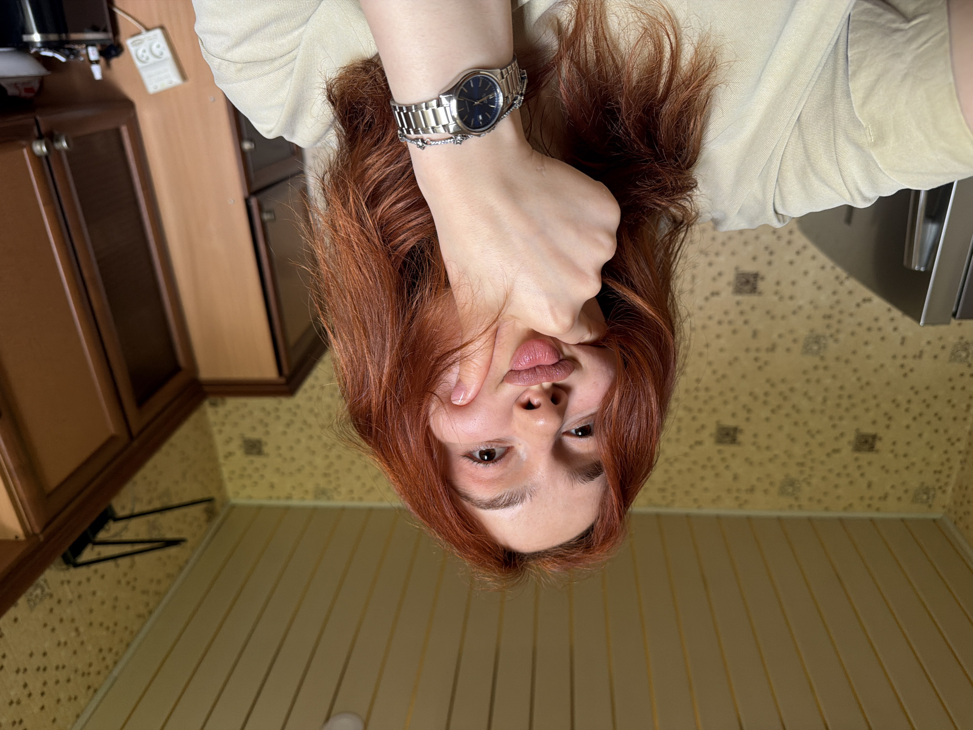 A woman with auburn hair is lying down, positioned so her face is oriented toward the bottom of the frame. Her left hand, adorned with a silver watch and bracelet, is raised and touching her face near her lips. She is wearing a light beige top. The background consists of a kitchen setting. There are brown cabinets and a white electrical outlet on the wall to the left. The floor is wood. The location cannot be determined from the image.