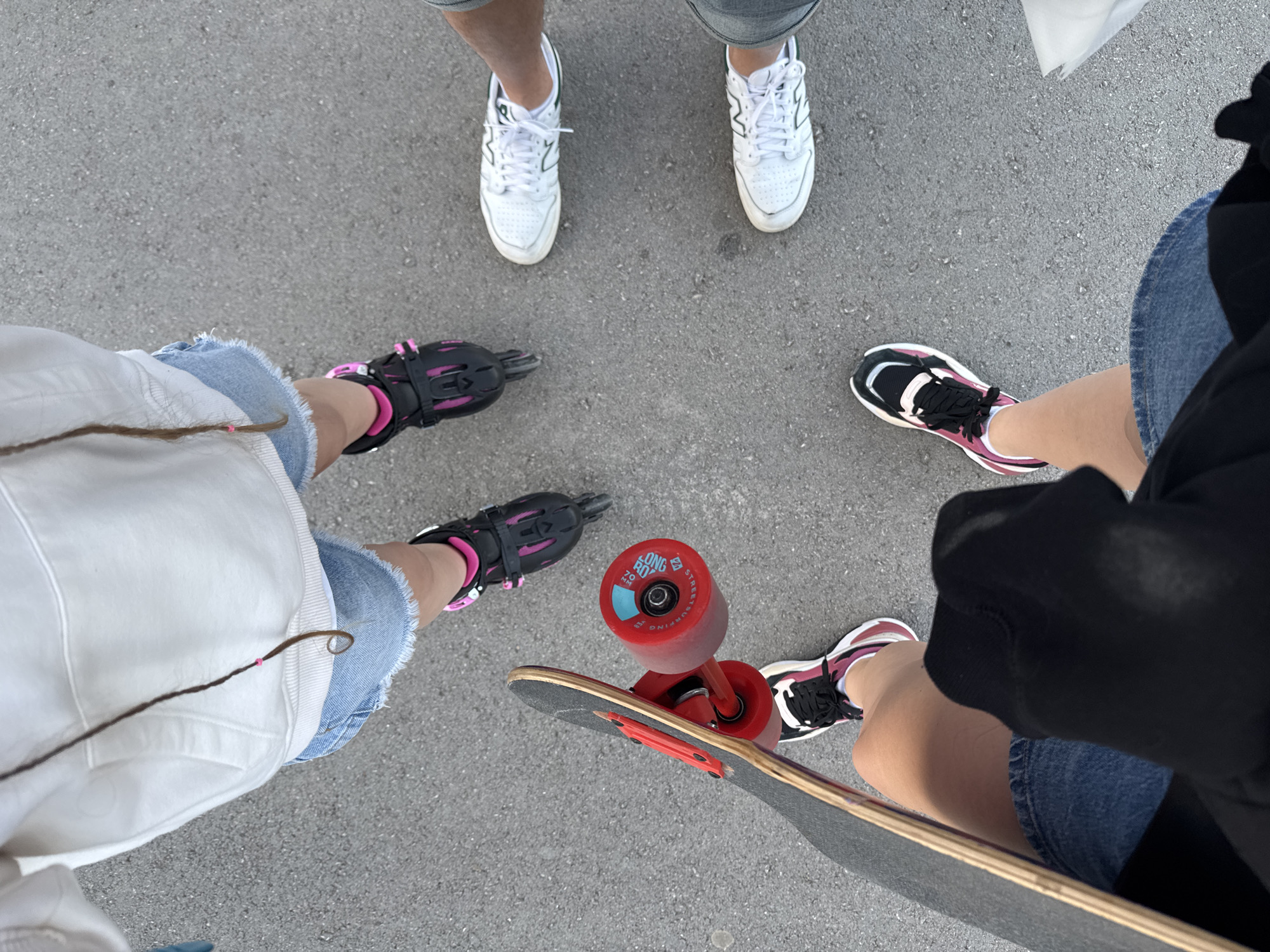 An overhead shot captures the lower bodies of four individuals standing on an asphalt surface in Budva, Montenegro. In the top-center, a person wears white New Balance sneakers and grey shorts. To the left, another individual is equipped with black and pink rollerblades, light blue denim cut-off denim shorts, and a white top, with a long brown braid featuring a pink bead hanging down. To the right, a third person wears black, pink, and white athletic sneakers, light blue denim shorts, and a black top, resting their hands on a skateboard. The skateboard has vibrant red wheels and trucks, with text "LONG BOARD", "STREET SURFING", "70mm", and "USA" visible on one wheel. A fourth person is partially visible at the top-right, wearing white sneakers and possibly holding a white bag.
The group appears to be gathered for outdoor recreational activities such as rollerblading and skateboarding. The setting is an open, paved area, likely a street or park. The time of day seems to be daytime, possibly late afternoon or early evening, characterized by soft, even lighting and a lack of harsh shadows. The overall mood is casual and relaxed, indicative of friends enjoying leisure time together.
