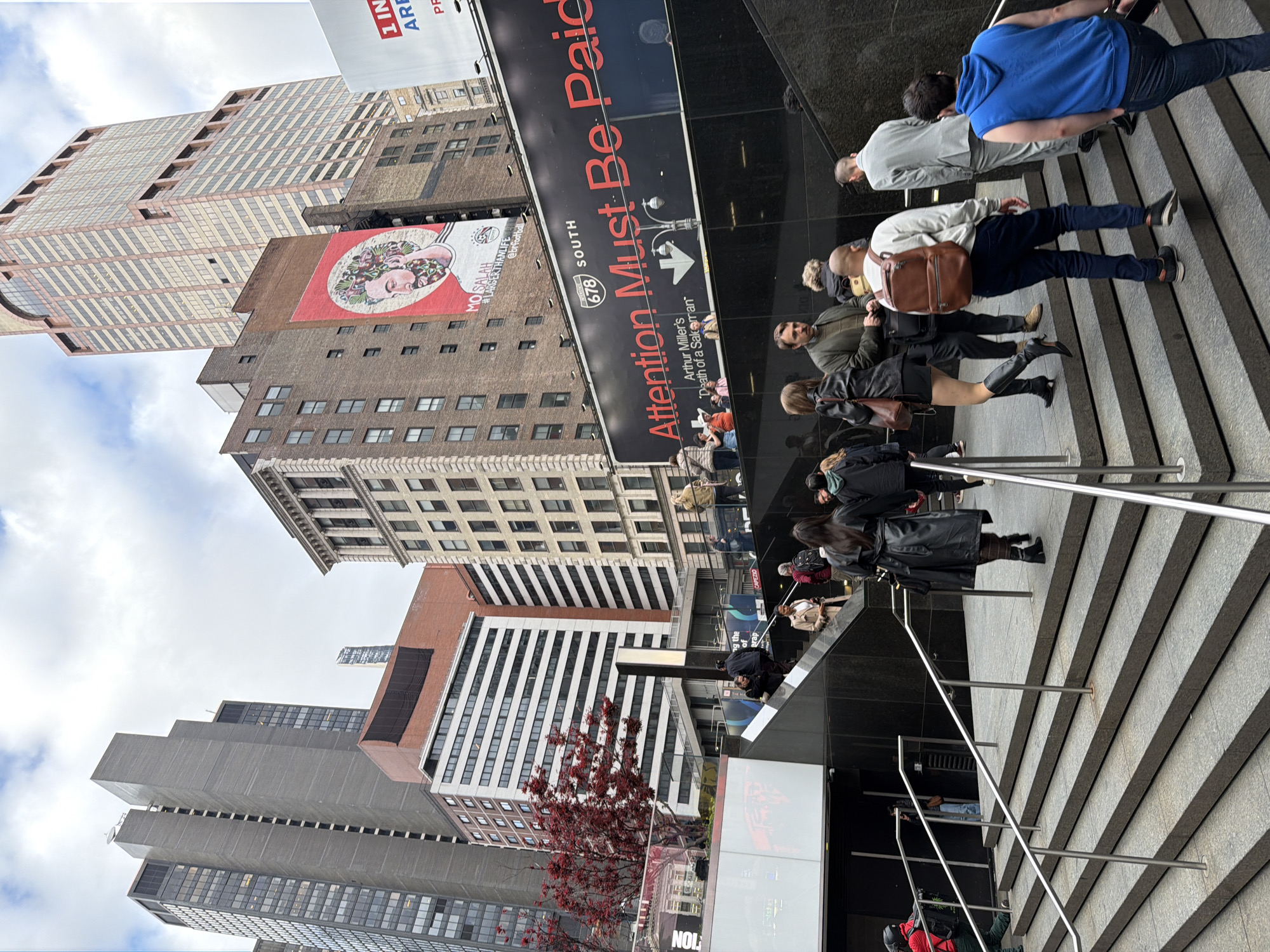 Here is a detailed description of the image:
The image captures a street scene in New York City, United States of America. The shot seems to be taken from below, looking up at the steps leading to a building. People are using the steps, both going up and down. The signage near the building indicates a play is showing, identified as Arthur Miller's "Death of a Salesman," with the phrase "Attention Must Be Paid." Nearby signage indicates that the location is Route 678 South.
Several buildings of varying heights and architectural styles surround the location. One building has a large advertisement or mural of what appears to be a celebrity figure. Other buildings have a modern design, with glass and steel structures. The sky above is partly cloudy. A few trees with red leaves are visible near the base of the steps. The light suggests it is daytime, but overcast.