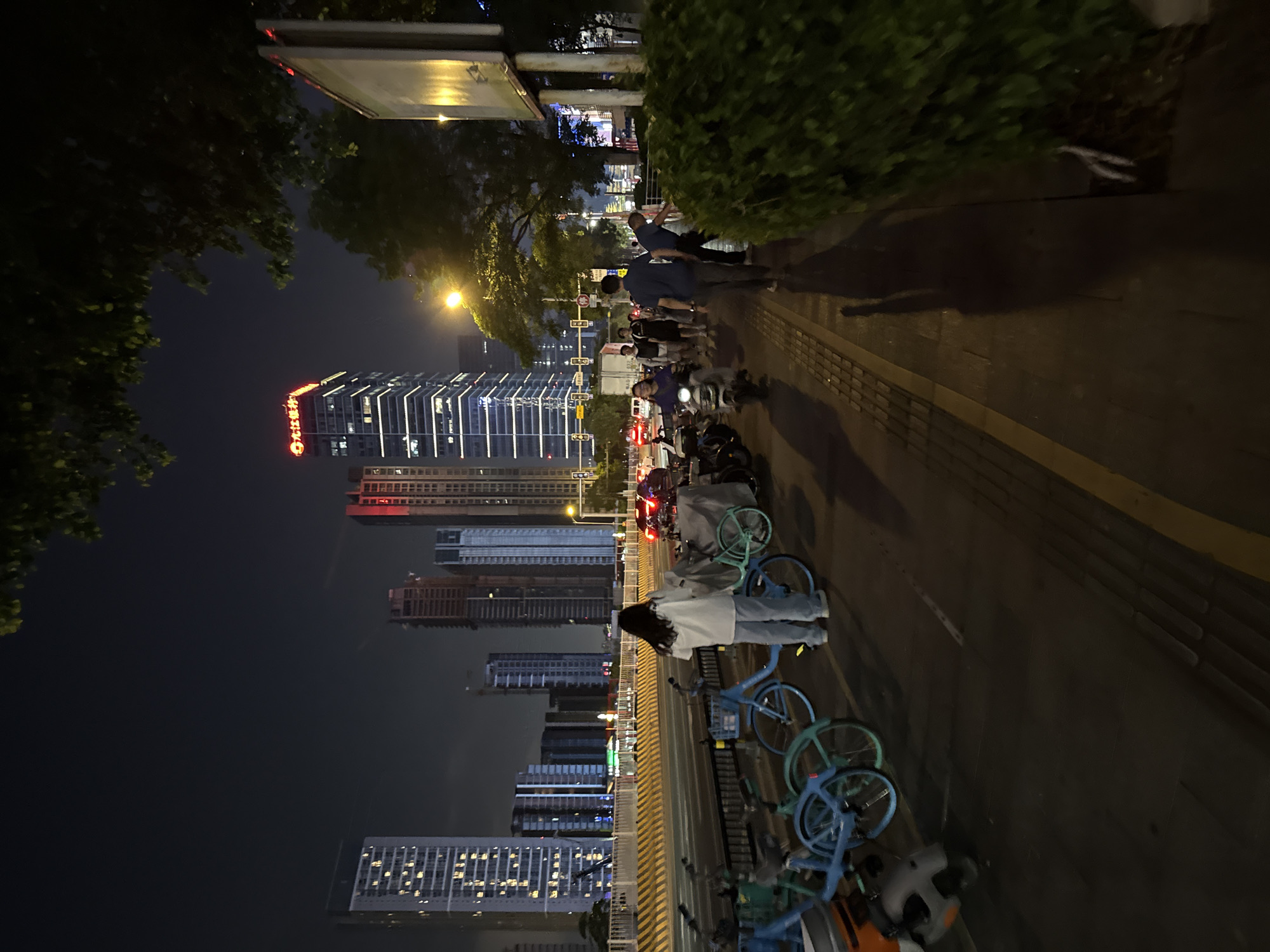 This is a nighttime street scene in Tianhe District, China. A woman stands on a sidewalk next to several blue shared bicycles. Behind her, the street is lined with yellow and black striped barriers, and beyond that, multiple high-rise buildings are visible. A red neon sign with Chinese characters that reads "大新银行" (Daxin Bank) is illuminated on one of the buildings. Further down the sidewalk, a group of people are walking, and a person is riding a scooter. Trees and bushes are present along the sidewalk. The street is illuminated by streetlights and the lights from the buildings.