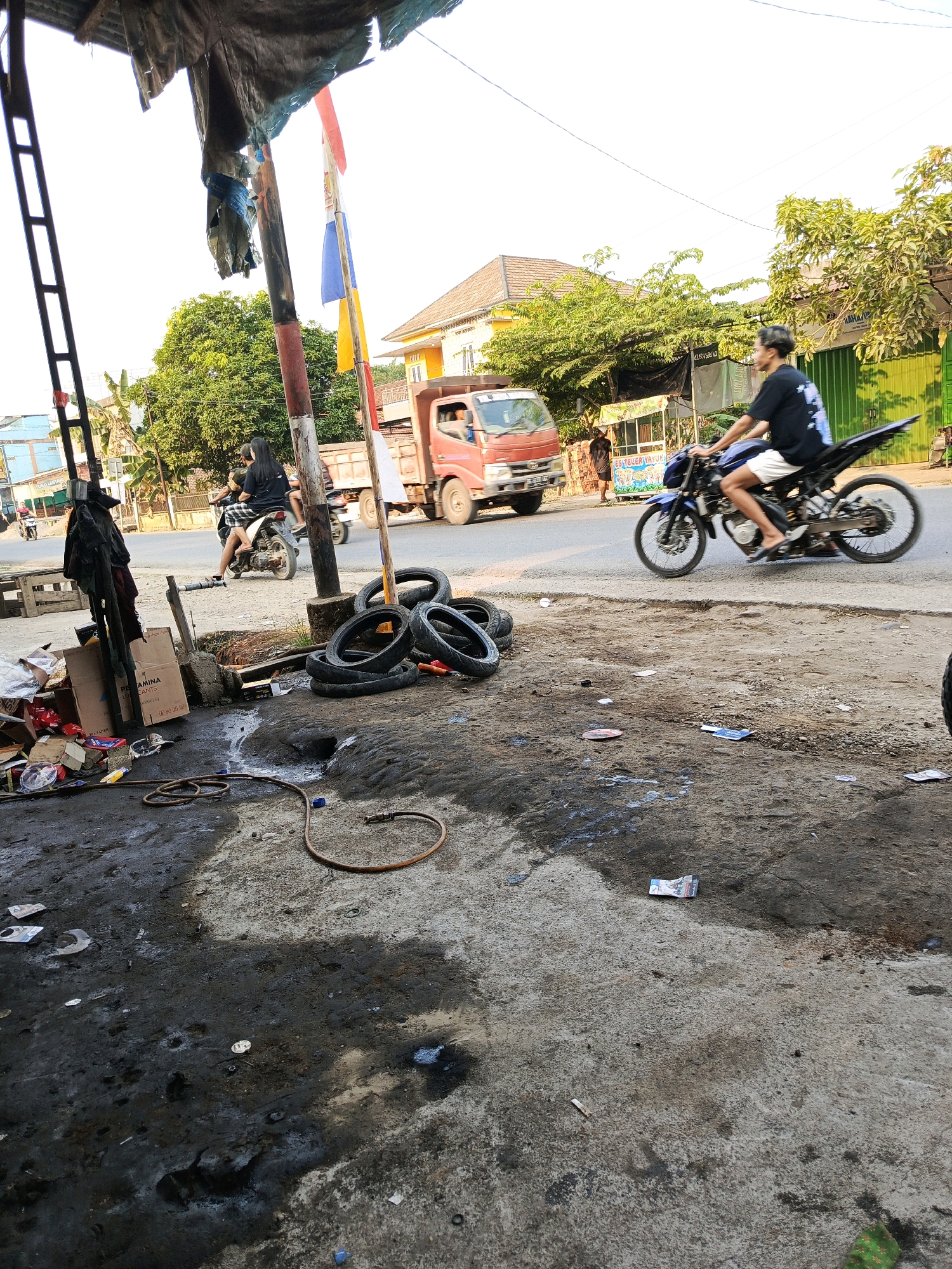 The image depicts a street scene in Megang Sakti I, Indonesia, with a focus on a roadside workshop. In the foreground, a wet, oily concrete surface is littered with debris, including old tires stacked near a rough-hewn wooden structure. A coiled orange hose lies on the ground. To the left, a pile of cardboard boxes and various items suggest a cluttered storage area.
In the mid-ground, a person rides a blue motorcycle with a black frame. They are wearing a black t-shirt and white shorts, with a tattoo visible on their left arm. The motorcycle has a distinctive rear design. Behind this, a woman with long dark hair is seated on a second motorcycle, wearing a checkered skirt and a black t-shirt.
Further in the background, a red truck is driving on the road. Various buildings and trees line the street, with colorful awnings visible on some structures. Indonesian flags, red and white, are flying from a pole. The overall impression is of a busy, somewhat disorganized, urban environment.