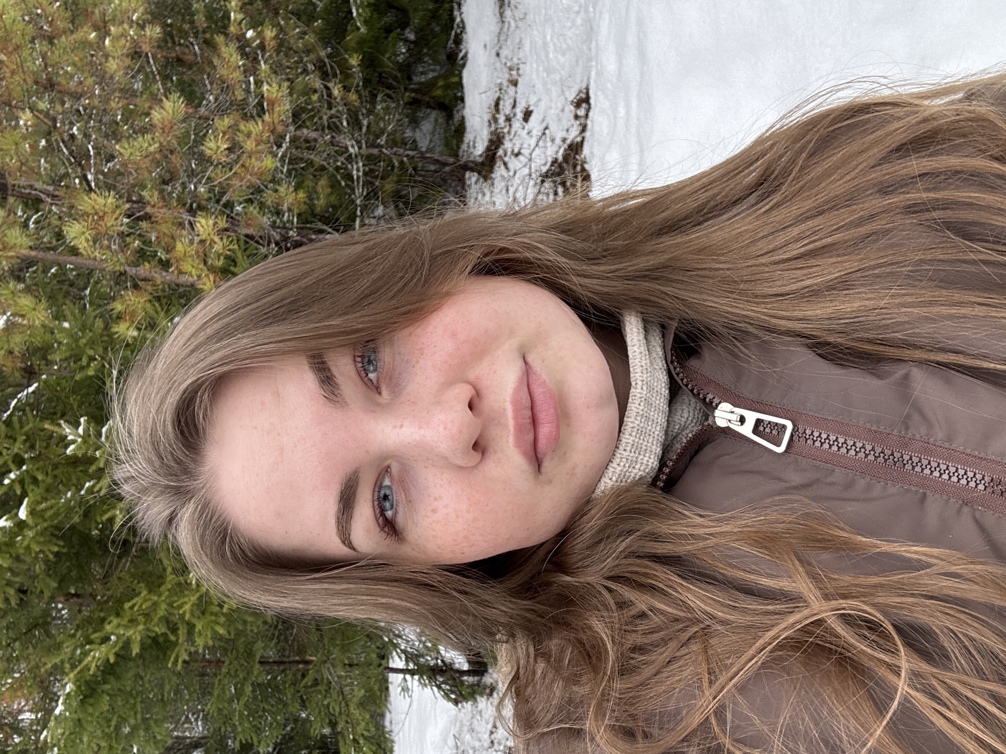 A young, fair-skinned woman with long, light brown hair is centered in the frame, slightly rotated. She has blue eyes and freckles on her face. She is wearing a brown jacket over a cream-colored turtleneck sweater. The jacket is partially zipped, exposing the zipper. Behind the woman is a natural environment with snow on the ground and evergreen trees. The specific location is Zavod Mikhailovskii, Russia. The woman appears to be taking a selfie.