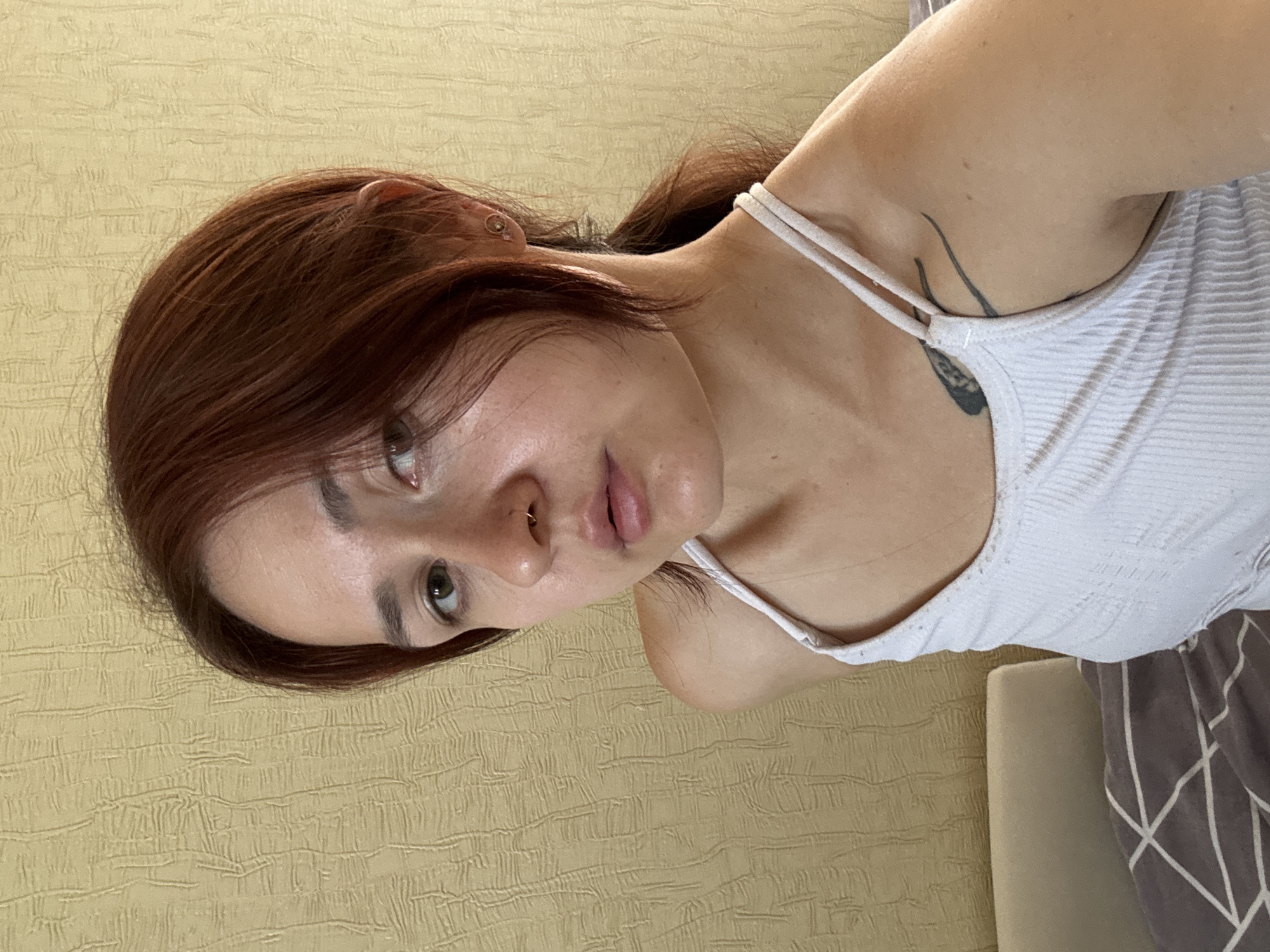A young woman with auburn hair pulled back into a ponytail is posing for a selfie in an indoor setting. She is wearing a white tank top and has a nose ring and an earring. A tattoo is visible on her left shoulder. The background appears to be a wall with a textured yellow wallpaper. The woman is positioned in front of a bed with grey and white patterned bedding. The lighting suggests it is daytime, likely indoors. There are no indications of the specific location within the city of Naberezhnye Chelny, Russia.
