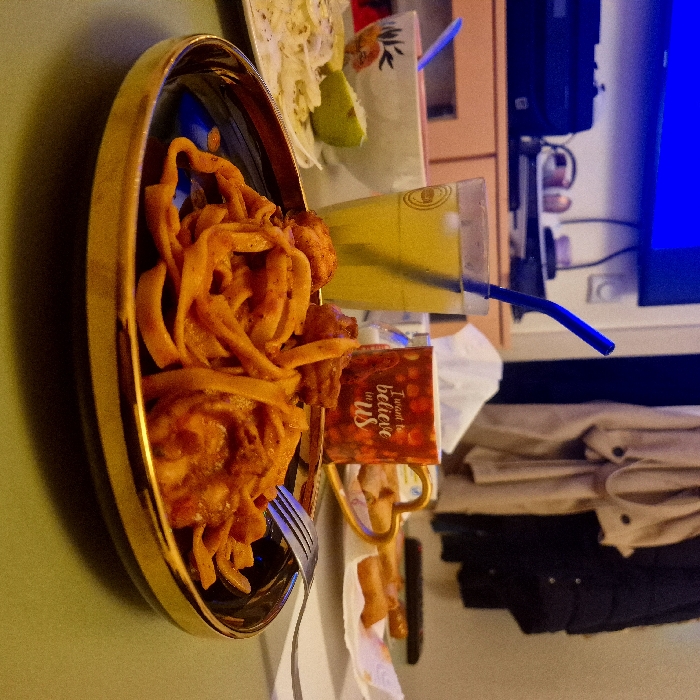 A golden oblong bowl contains a heap of orange, curled snacks next to a salad. A tall glass of yellow liquid with a blue straw is placed beside it. A small sign reading “I want to believe in us” rests by the bowl. A fork sits on the table, and the background features a TV screen and stacked jackets.