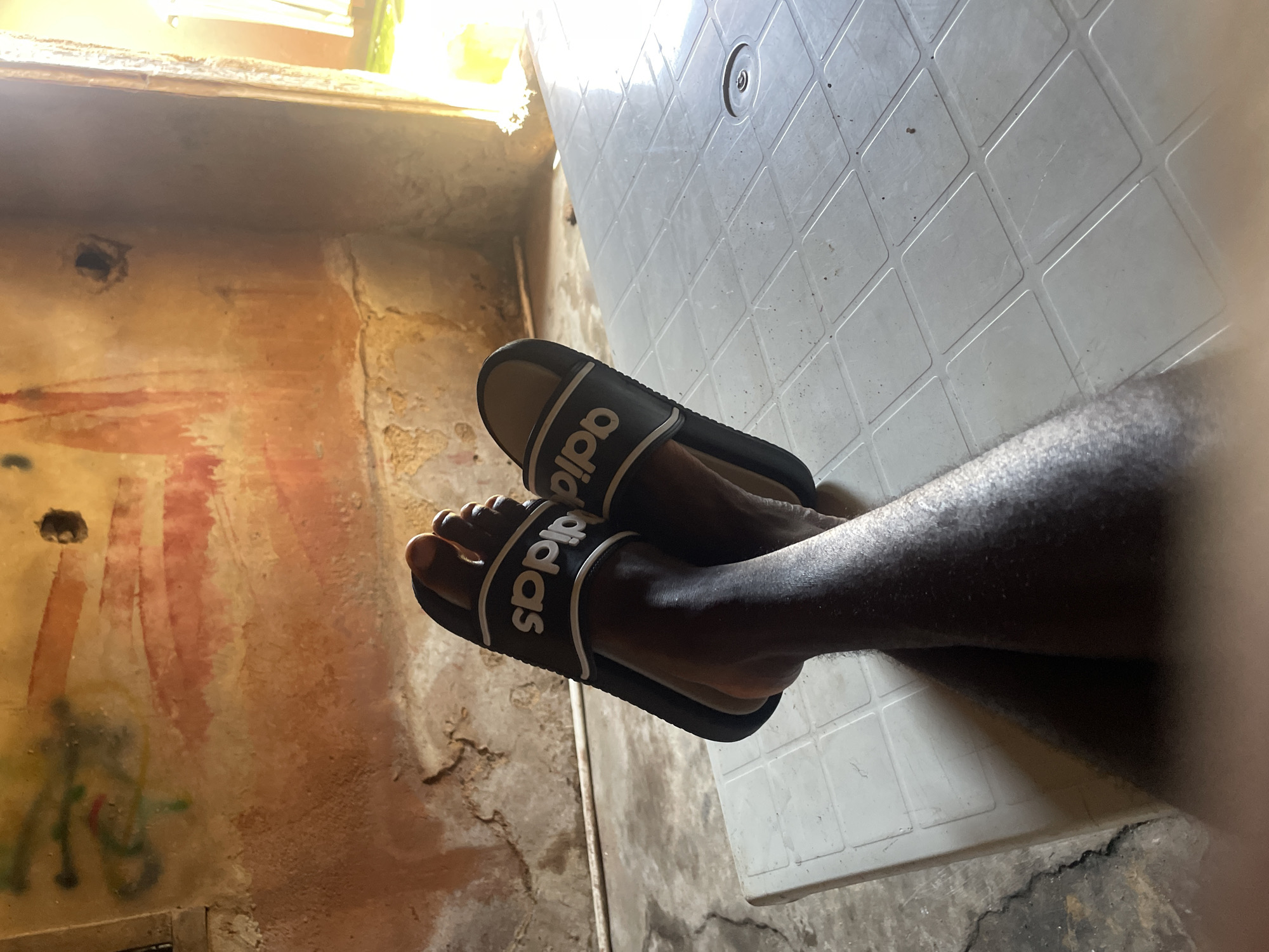 This is a close-up, eye-level shot of a person's legs and feet wearing black Adidas slides, resting on a white plastic table. The background consists of a wall with peeling paint, showing shades of orange, red, and yellow, with some green spray paint graffiti. There is a visible hole in the wall, and a pipe runs horizontally along the lower part of the wall. To the right, an opening reveals a brightly lit outdoor space. The scene appears to be indoors, possibly in a residential or informal setting in Jos, Nigeria. The lighting suggests it is daytime.