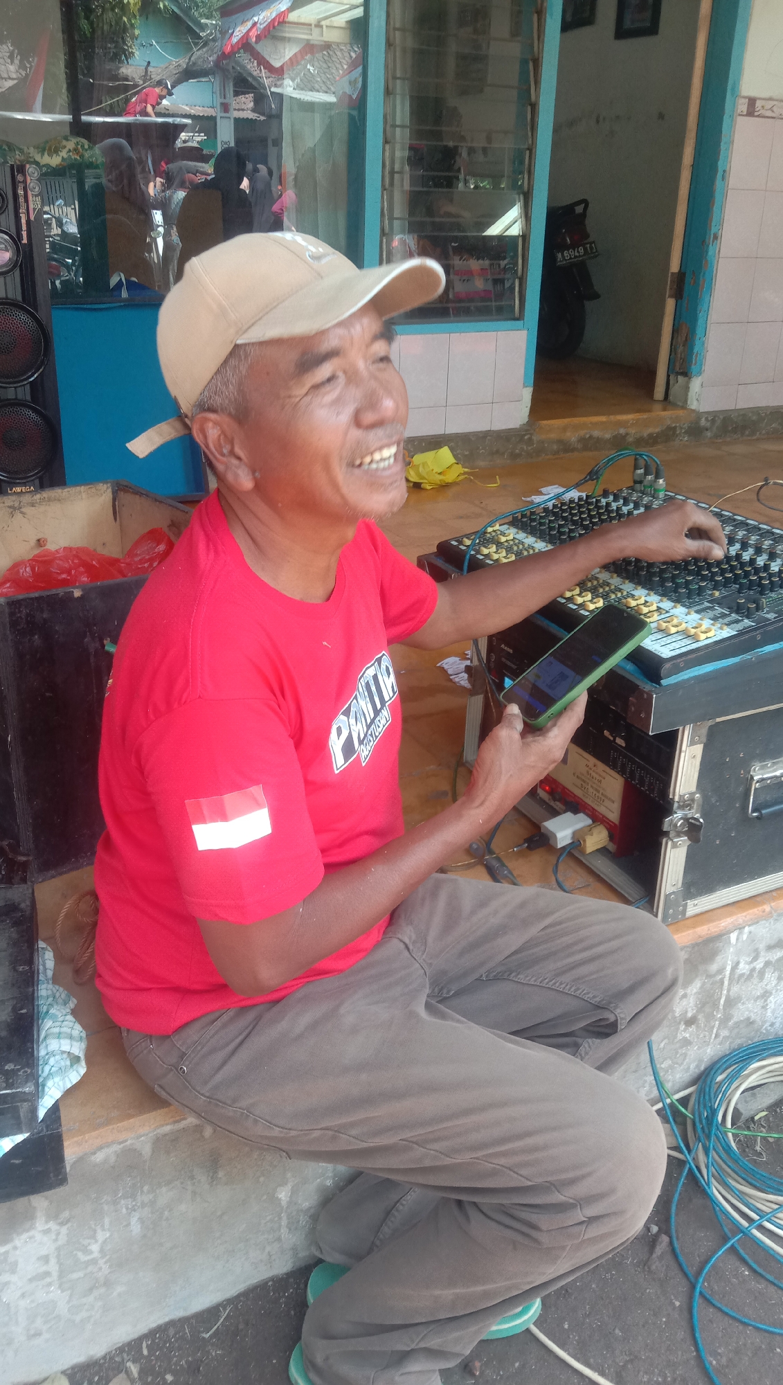 In the city of Ngempit, Indonesia, an elderly Indonesian man wearing a red t-shirt with a white and red flag patch on his left sleeve and a tan baseball cap is seated on a concrete ledge outdoors. He is holding a green smartphone in his right hand and adjusting knobs on a black audio mixer with his left hand. The audio mixer is placed on top of a black equipment case. In the background, a building with blue trim and a window with bars is visible. A motorcycle with a license plate reading "N 6949 TJ" is parked inside the building through the window. Various audio equipment, including speakers and cables, are also present in the scene. The ground is made of concrete and dirt.