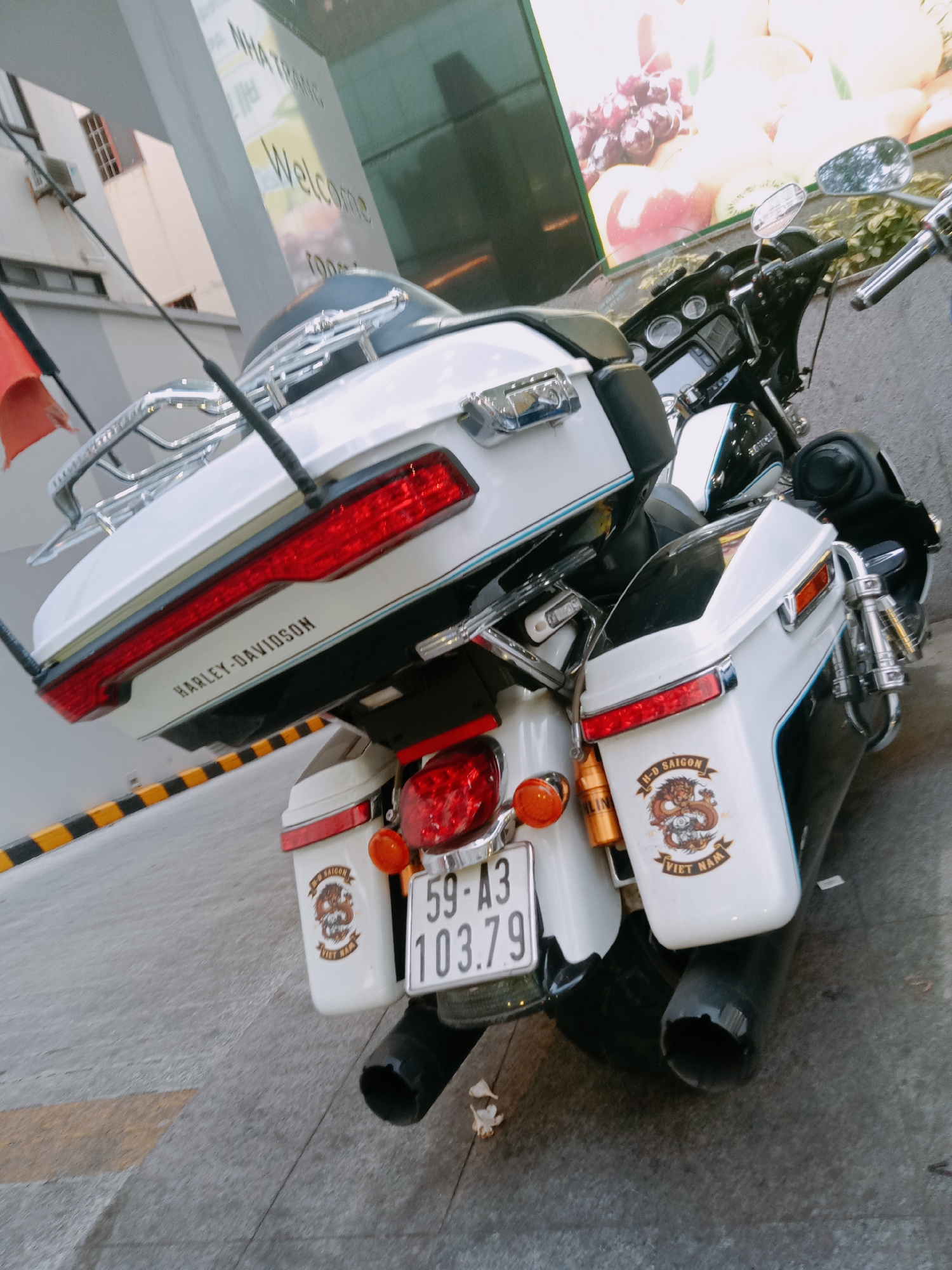 A slightly tilted eye-level outdoor shot depicts a white Harley-Davidson motorcycle parked on the sidewalk. The motorcycle is adorned with various attachments, including a chrome luggage rack and a prominent tail light. The tail features “HARLEY-DAVIDSON” inscribed in black lettering. Distinct white saddlebags are mounted on either side of the rear wheel, each adorned with a brown and white emblem with the words "H-D SAIGON VIET NAM" . The motorcycle license plate is visible and reads "59-A3 103.79".
In the background, a building is partially visible, sporting a sign reading "NHA TRANG" and "Welcome". The scene is set in Nha Trang Ward, Vietnam. A close look shows a wall-sized image of fruits and vegetables, possibly an advertisement, suggesting the building may be related to food or produce.