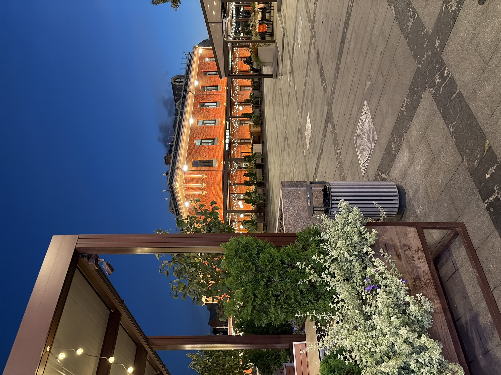 A street scene in Moscow, Russia, at dusk. A red brick building with illuminated windows and decorative lights stands across a paved courtyard. To the left, a covered seating area with string lights and potted plants is visible, with a wooden planter filled with greenery in the foreground. A trash receptacle is positioned near the planter. The sky is a deep blue, indicating twilight.
