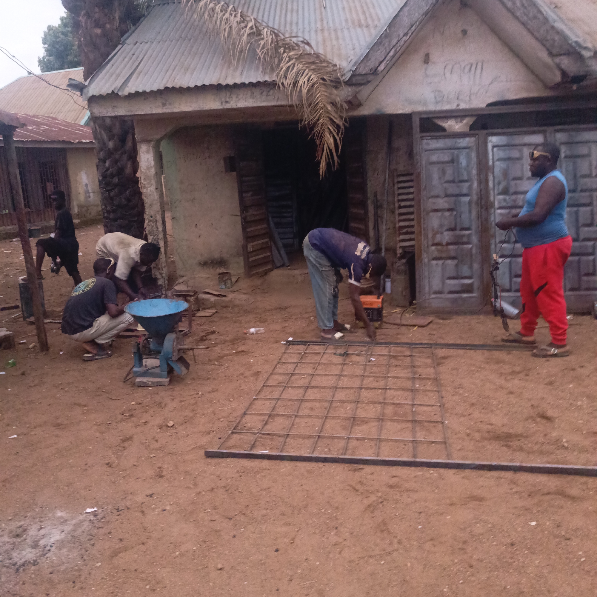 The image depicts a scene in Akwanga, Nigeria, where a group of men are working on a metal structure. One man, wearing a blue tank top and red pants, stands with welding equipment near a metal grid on the ground. Another man in a blue shirt is welding the grid. Two other men are working on a blue machine that looks like a grinder. A fifth man sits in the background. The work area is outside a building with a corrugated metal roof and a large double door. The ground is bare earth.