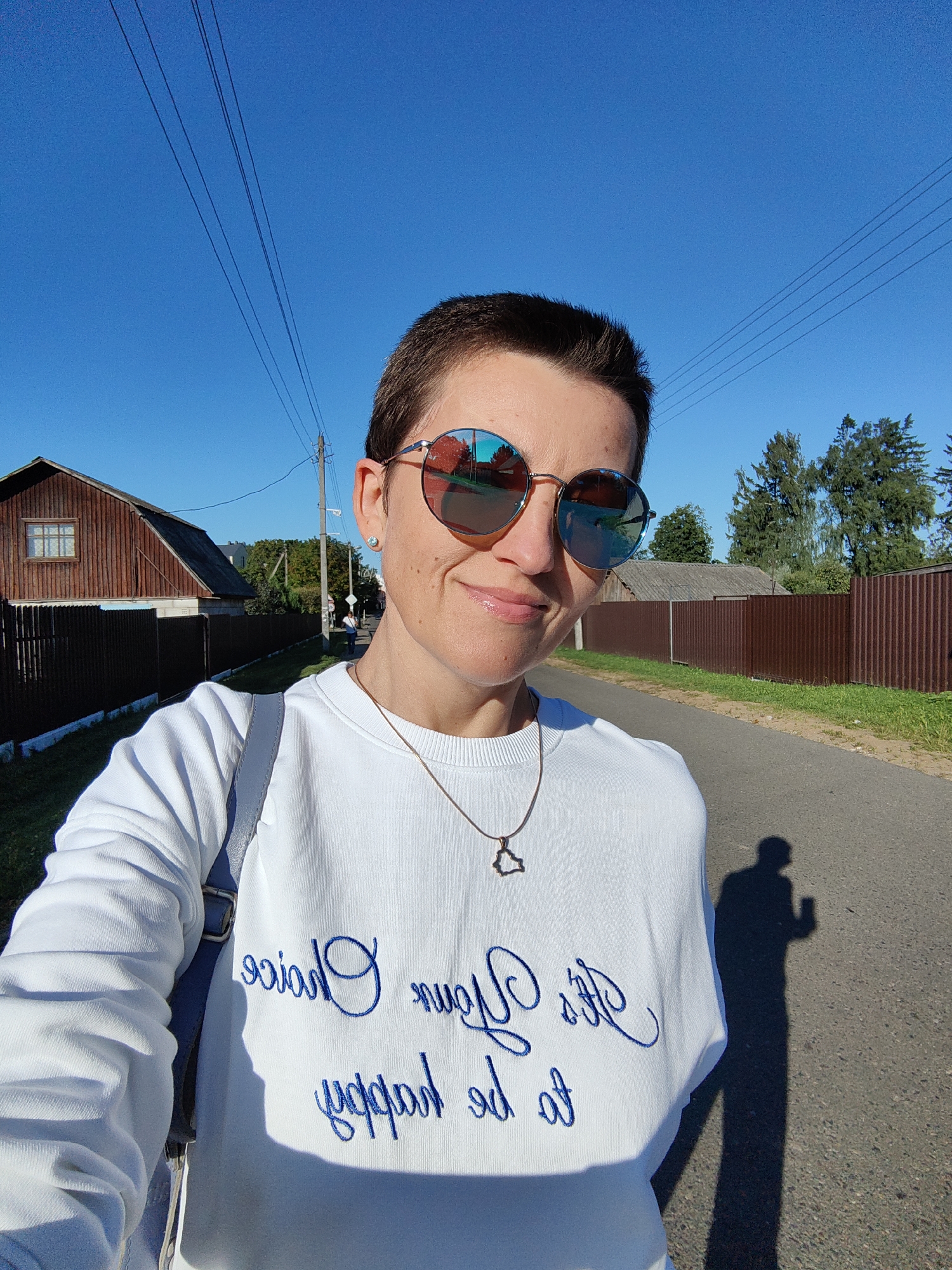 A woman with short dark hair and sunglasses takes a selfie outdoors in Fanipal, Belarus. She is wearing a white sweatshirt with blue cursive lettering that appears to read "Your choice to be happy." A gray strap from a bag is visible over her shoulder. She is standing on a paved road in front of a wooden house with a brown fence. Trees and a clear blue sky are in the background. A shadow of the woman is cast on the road to her right.