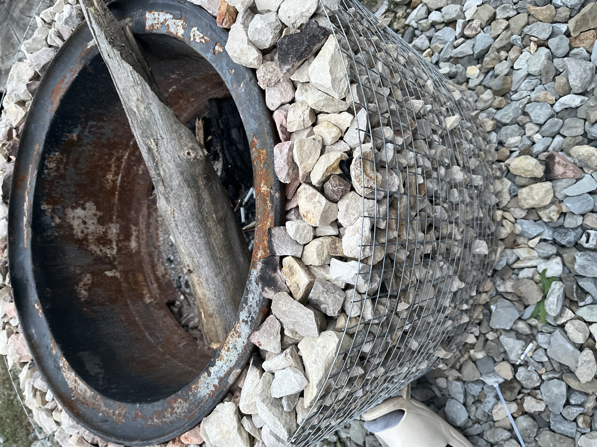A fire pit constructed from a repurposed metal drum, lined with rocks, sits outdoors. The drum's interior shows signs of rust and charring, with a piece of wood resting inside. The exterior is partially encased in a wire mesh filled with light-colored gravel, and more gravel surrounds the base. A single light-colored slipper is visible to the left of the fire pit. The ground is covered in small, grey and beige stones, with a few blades of grass. There is no indication of the specific location within Saratov, Russia.