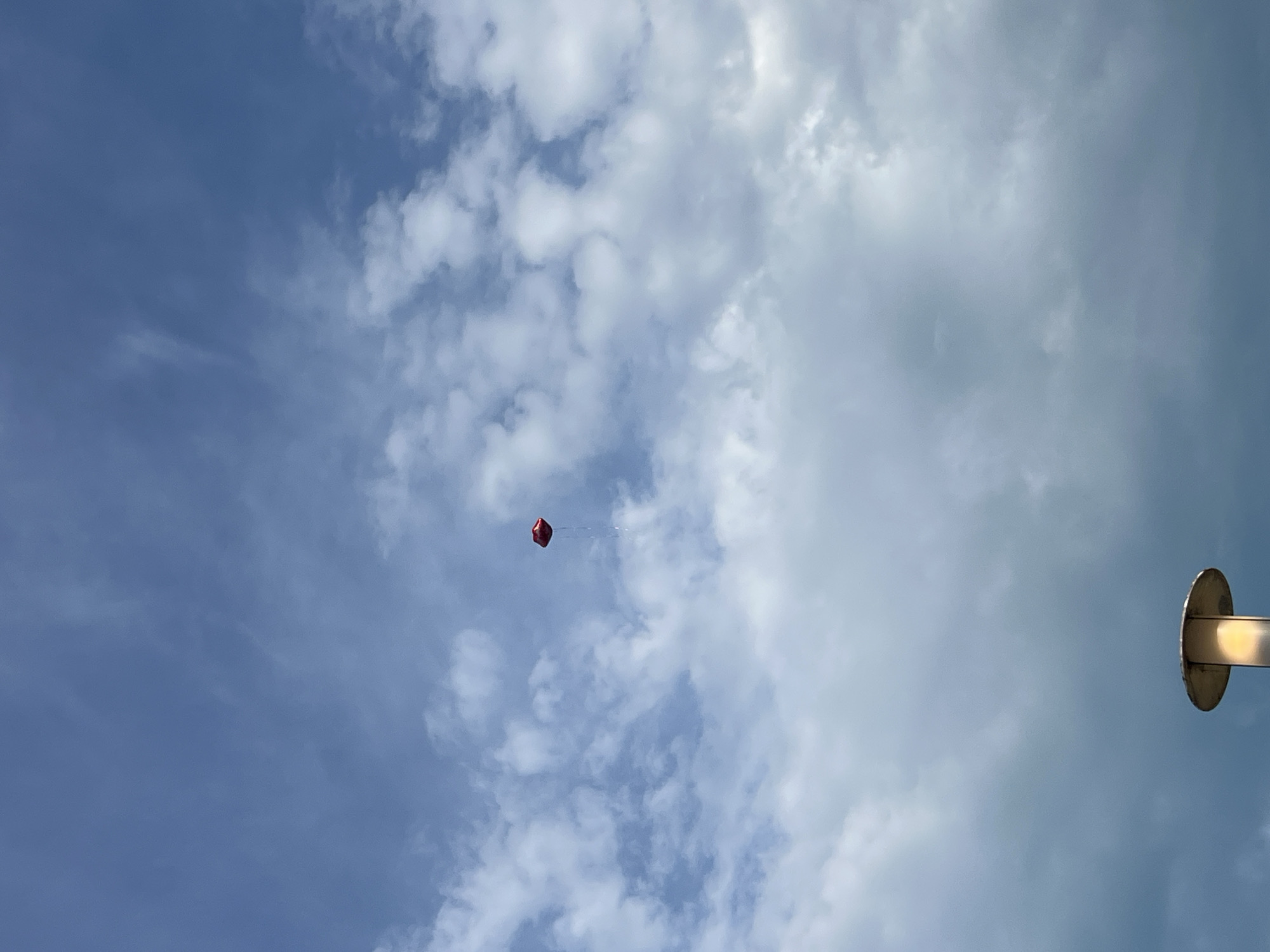 The image displays a bright, partly cloudy sky during daytime. The sky on the left side of the frame is a clear, deep blue with wispy white clouds, while towards the right, the clouds become denser and more uniform, creating a lighter, almost overcast appearance. In the central-left portion of the frame, a single, vibrant red balloon floats, appearing somewhat angular or diamond-shaped due to its inflation and perspective, with a faint string or tether trailing below it. Along the far right edge of the photograph, a metallic, circular disc is partially visible, attached to a pole or arm that extends horizontally inwards from the frame's edge. This could be part of a lamppost or a similar overhead fixture. No activity other than the balloon floating is discernible, and there is no visible text. The location context provided is Varna, Bulgaria.