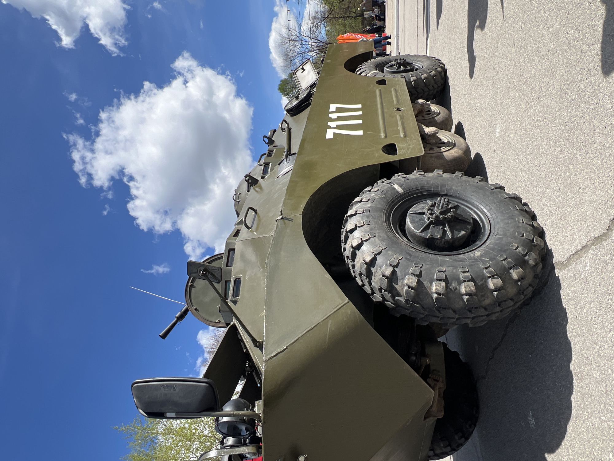 A green armored personnel carrier, identified by the white number "7117" painted on its side, is parked on an asphalt surface under a partly cloudy blue sky. The vehicle has large, knobby tires and is equipped with what appears to be a machine gun mounted on top. The location is potentially Cherepovets, Russia. The presence of trees and indistinct figures in the background suggests an outdoor setting.