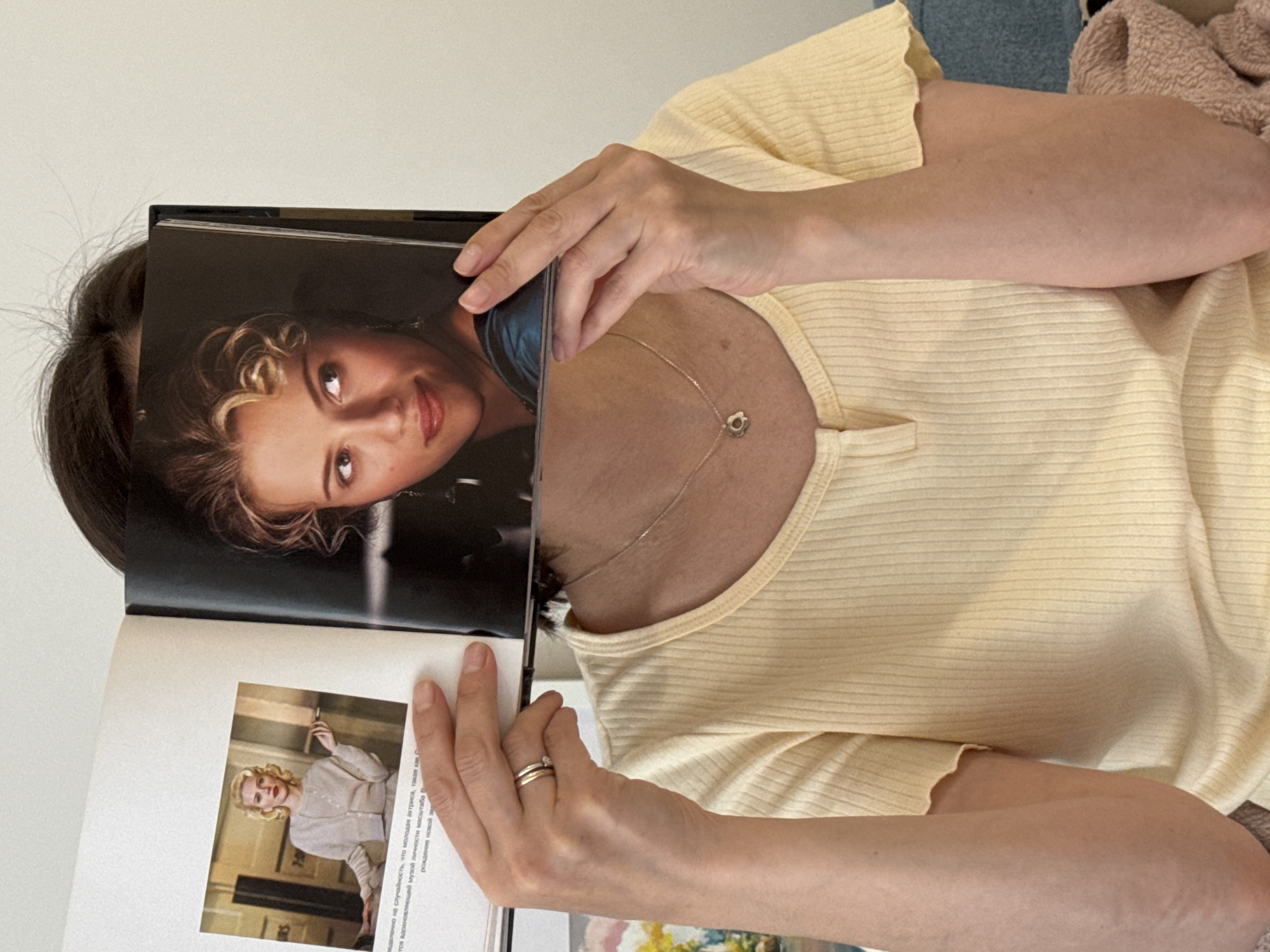 A woman is holding an open book in front of her face. The book has a photograph of a woman on the left page. The photograph is a portrait of a blonde woman with her hair styled in curls and the woman's head is visible over the upper right corner of the book. The woman holding the book is wearing a yellow, short-sleeved shirt and a necklace with a small charm. The pages of the book are printed with text in Russian. The text on the right page reads: "ределенно не случайность, что молодая актриса, такая как С тся вдохновляющей музой личности масатаба В рождение новой зе". The background is a plain white wall. The woman is also wearing a ring on her ring finger. Part of another person is shown in the upper right corner of the image. The person is wearing blue jean shorts. No location data is available for the city of Izhevsk, Russia.