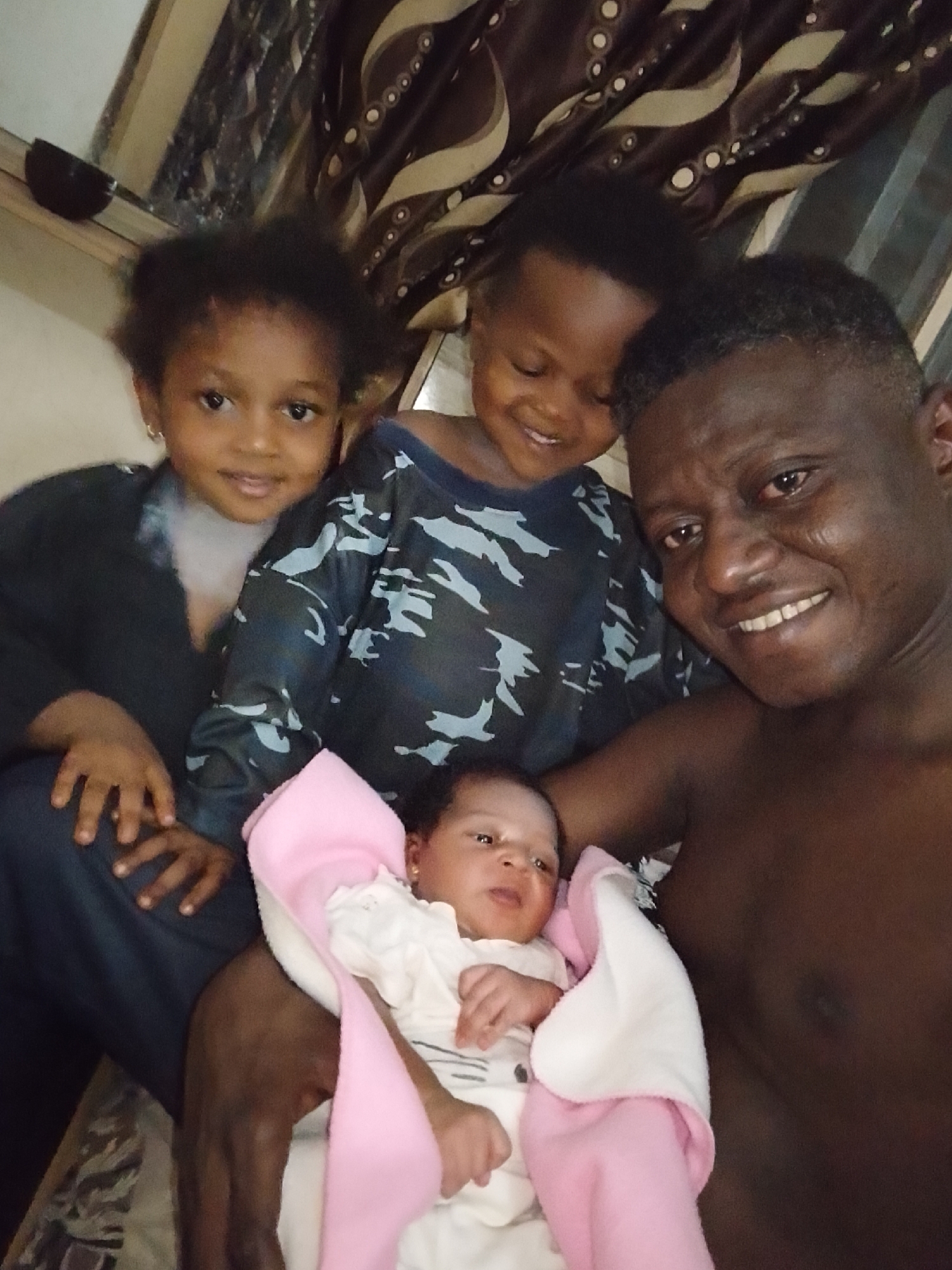This media file captures a close-up indoor family portrait featuring a dark-skinned man and three children, likely in Yelwa, Nigeria. The man, positioned on the right and center, is shirtless, sporting a short, dark haircut, and a broad, warm smile directed at the camera. He appears to be holding a newborn infant, who is wrapped in a soft pink and white blanket and wears a white outfit. The infant has an alert expression and looks slightly upwards.
To the man's left, a young girl with dark, curly hair and hoop earrings smiles directly at the camera. She is wearing a dark top. Partially visible behind her and above the infant is another young child, a boy, with short dark hair, who is smiling with his gaze directed slightly downwards. He is wearing a long-sleeved shirt with a camouflage-like pattern in dark blue and light blue hues.
The group is tightly framed, suggesting a selfie or a very close-up shot. The background reveals patterned curtains with shades of brown and beige, and a glimpse of an interior wall, indicating an indoor setting, possibly a home. A dark bowl or object is visible in the top left corner. The lighting is soft, consistent with an indoor environment. The overall mood is one of happiness and familial affection. There is no visible text in the image.