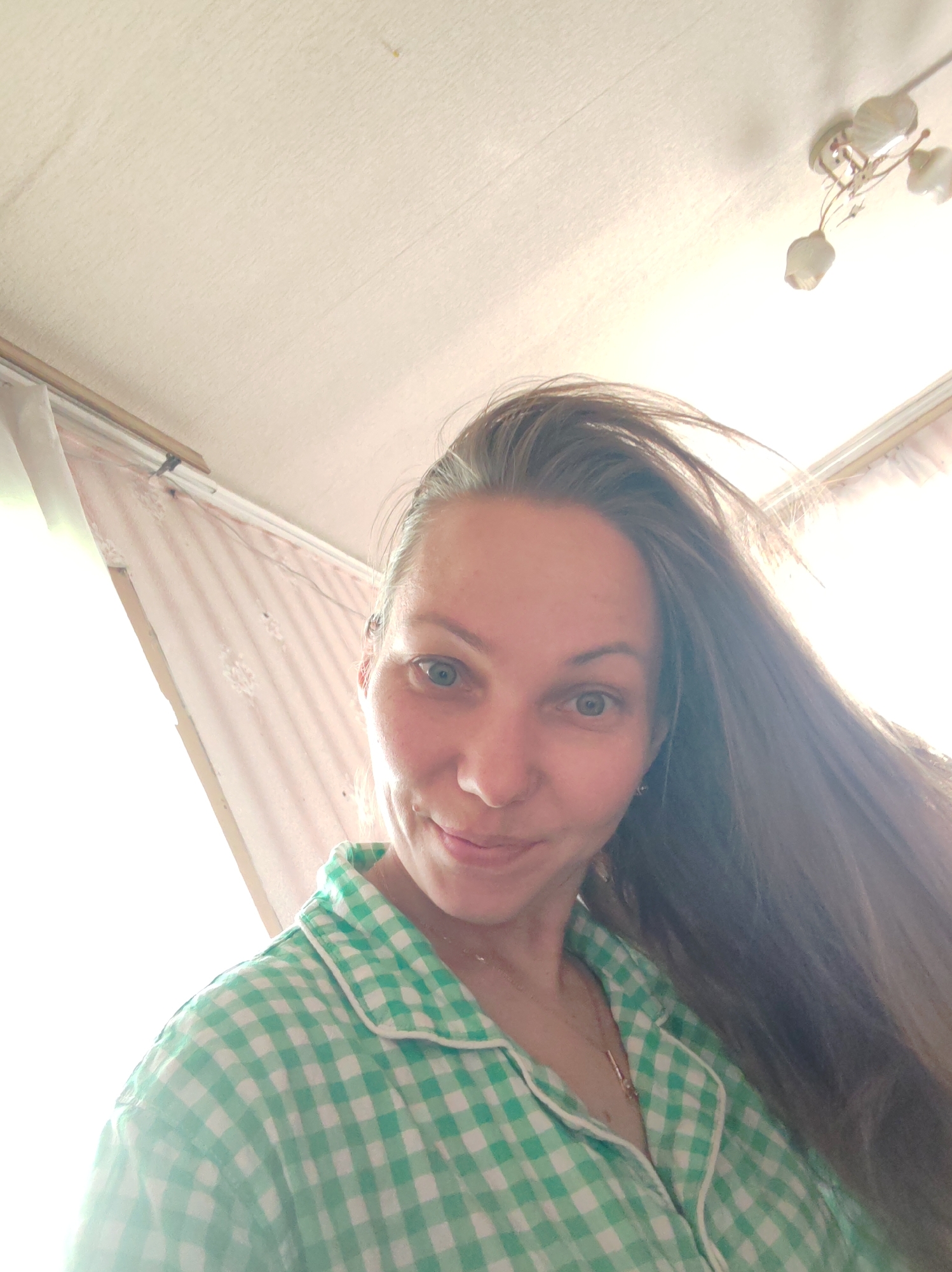 A fair-skinned woman with long brown hair, blue eyes, and a slight smile is depicted in a selfie. She wears a green and white checkered pajama top. Behind her, a room is visible with a white ceiling, a light fixture, and a pink and white patterned curtain. The image appears to be taken indoors. There is no information about the city of Чухлэм, Russia within this image.