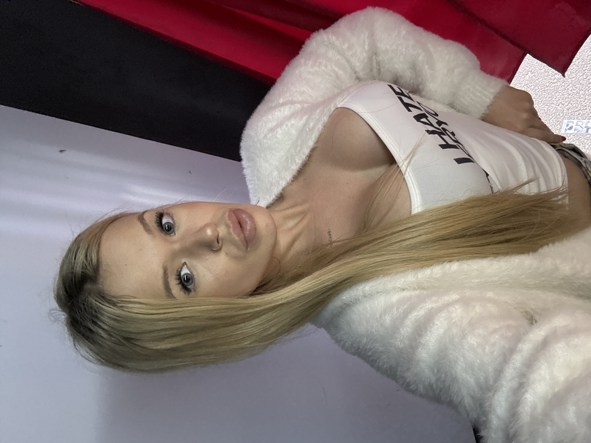 A blonde woman with blue eyes and full lips is taking a selfie. She is wearing a white tank top with the words "I HATE EVERYONE" printed on it, and a fluffy white cardigan. She has a nose ring and a small tattoo on her neck. The background appears to be a wall with vertical stripes of white, black, and red. There is no indication of the location in Saint Petersburg, Russia.