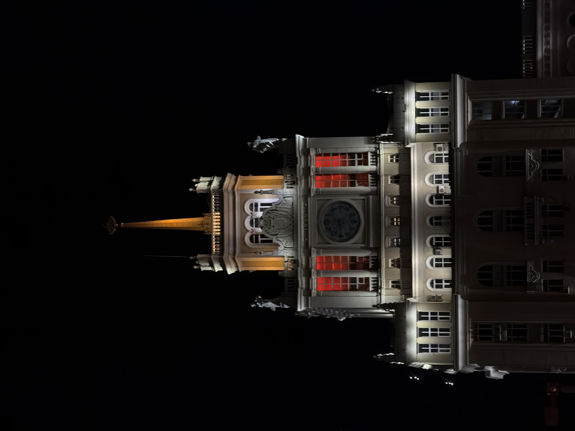 The image shows the Kotelnicheskaya Embankment Building, one of the Seven Sisters skyscrapers in Moscow, Russia. It is a nighttime shot, with the building illuminated against a dark sky. The architectural details are visible, including the spire, clock, sculptures, and windows. Some windows are lit with red light. The building is viewed from a low angle.