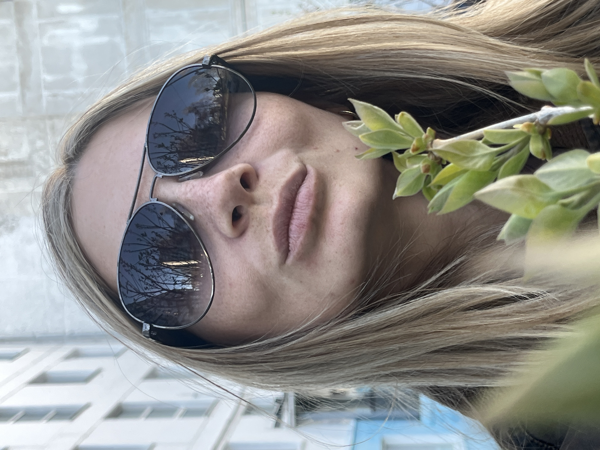 A close-up, tilted portrait photograph depicts a fair-skinned woman with long, blonde hair. She is wearing aviator-style sunglasses that reflect a cityscape with bare tree branches against a bright sky. A branch with green leaves is positioned near her face on the right side of the frame. The background includes a building with white-framed windows and blue accents, suggestive of an urban setting. Given the location of Chaikovsky, Russia, and the reflection in the glasses showing a tree with bare branches, it is likely the image was taken in the early spring when trees haven't fully grown their leaves.