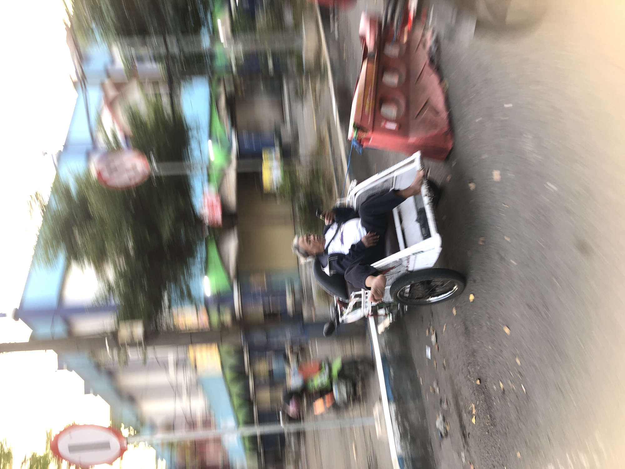 The image shows a man sitting in a motorized rickshaw, looking at his phone. The rickshaw is white with silver accents and a black seat. The man is wearing a white shirt with a dark jacket and dark pants. His feet are bare. The rickshaw appears to be in motion, as evidenced by the motion blur in the image.
In the background, there is a building with blue and white trim and a green awning. There are trees and street signs visible, including a red and white circular sign with a white vertical bar. A green motorcycle with an orange seat is parked behind the rickshaw. A red traffic barrier is also visible in the foreground, to the right of the rickshaw. The scene is outdoors, on a paved street, with debris on the ground. The overall impression is of a busy urban environment. The context provided indicates that this scene is located in Larangan, Indonesia.