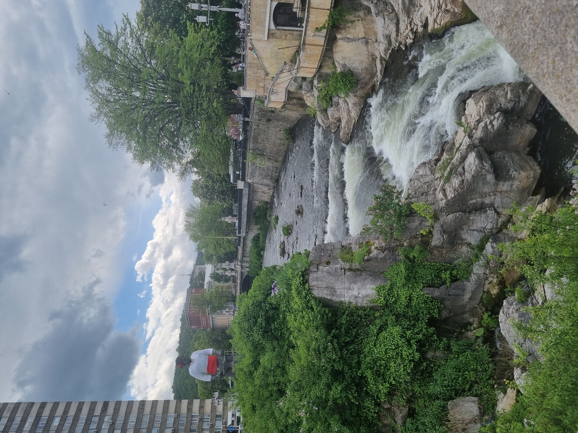 The image shows a view of the Yantra River in Gabrovo, Bulgaria, looking downstream towards a small waterfall or rapids. The river flows between rocky banks covered in vegetation. On the left bank, there is a stone embankment with buildings and trees. A statue of a man in white shirt and a red sash is seen to the left of the frame. A modern, tall building is seen at the bottom left. The sky is partially cloudy with patches of blue. The water is rushing and white where it falls over the rocks. The light suggests it is daytime. A bridge or other structure is partially visible on the right side of the frame.