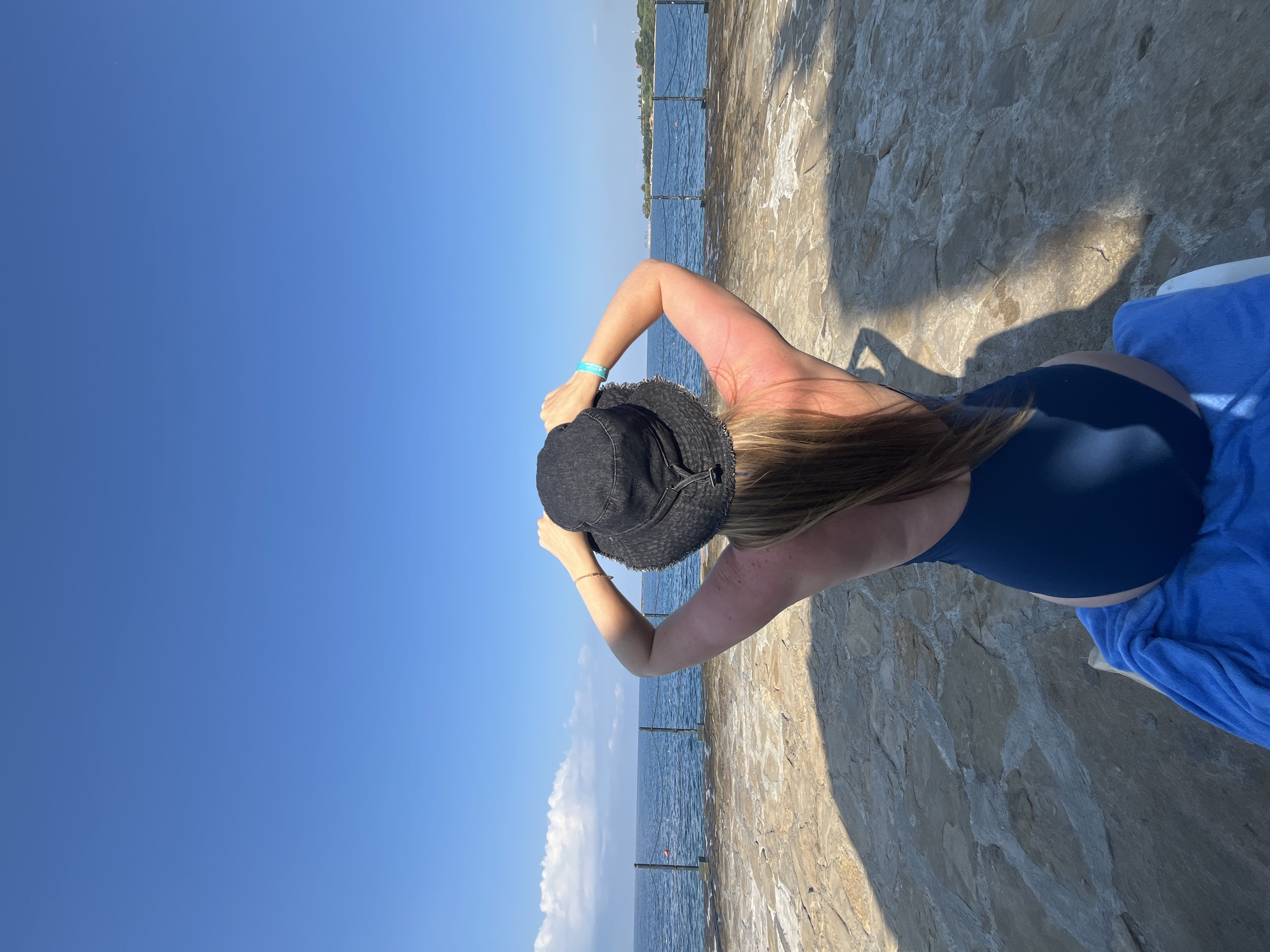 A woman wearing a dark blue one-piece swimsuit and a black wide-brimmed hat is positioned in the foreground, facing away from the camera. She is seated on a blue towel, with her hands on top of her hat, adjusting it. Her back and shoulders are visible, and she appears to have a tan. She is looking out towards a body of water, likely the Mediterranean Sea, with a clear blue sky above. In the middle ground, a stone pathway leads to the water's edge, where a metal railing is installed. Beyond the railing, the sea stretches out, with a distant shoreline visible on the right, featuring some buildings and greenery. The sun is casting shadows across the stone pathway. This scene is consistent with the coastal landscape found in Alanya, Turkey.