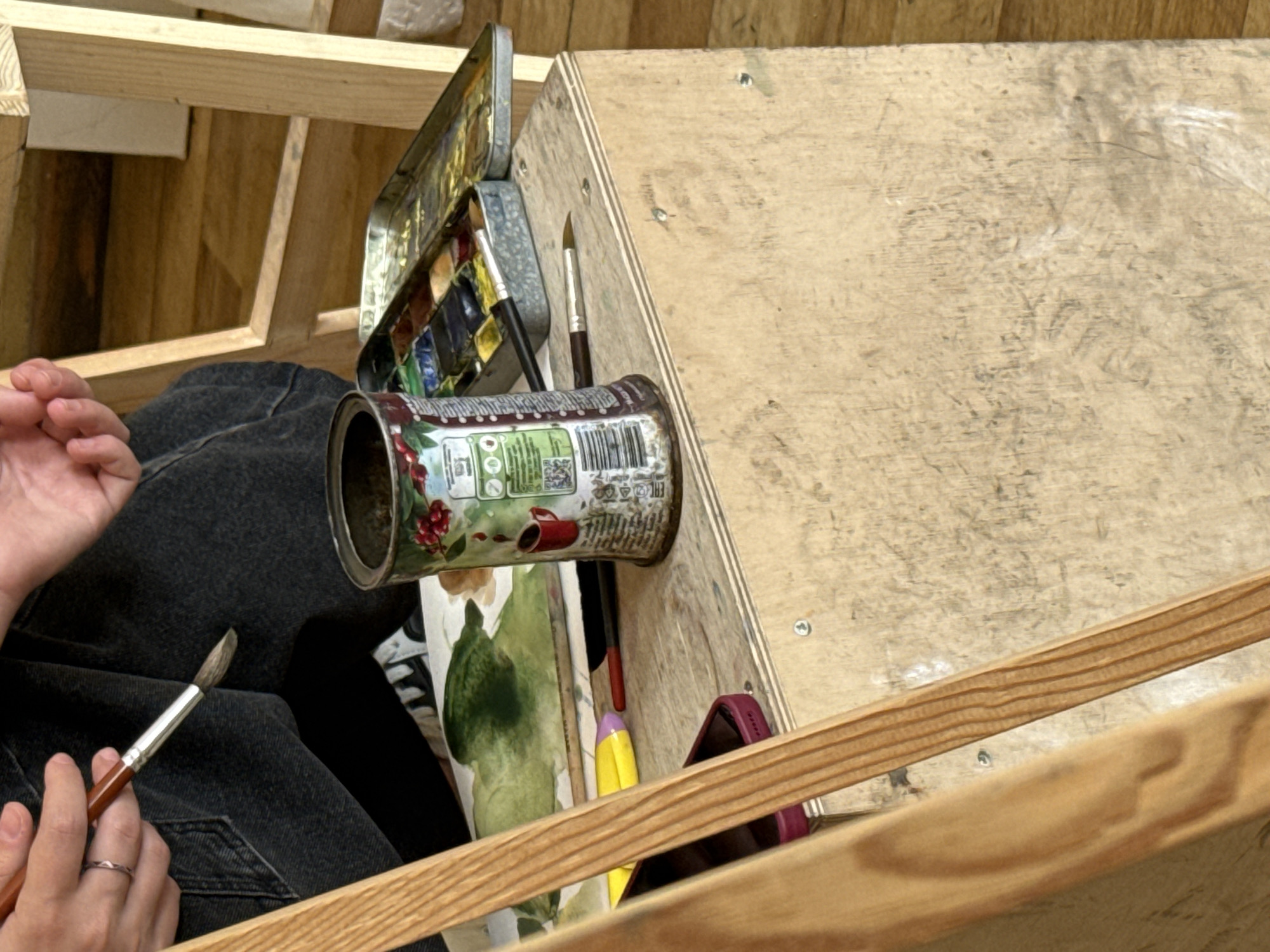 A high-angle shot shows a person seated in front of a wooden artist's easel, likely indoors. The person wears dark jeans and holds a paintbrush in their left hand, with a silver ring visible on the ring finger. An open paint palette with various colors sits to the left of the easel. A recycled tin can, possibly repurposed as a water container or brush holder, rests near the palette. Several other paintbrushes are scattered nearby. A painting in progress, featuring green and white strokes, is visible on the easel. The setting appears to be an artist's studio or workspace. Yerevan, Armenia is not indicated in the image.