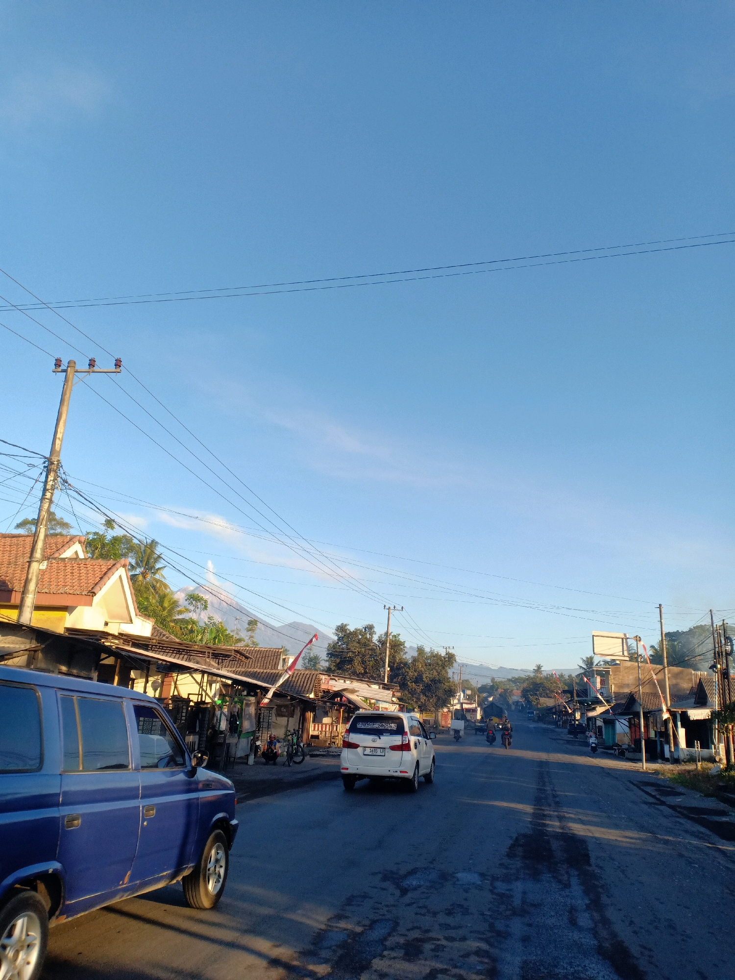 The image depicts a street scene in Pasirian, Indonesia, under a clear blue sky. On the left side of the road, a blue SUV is parked facing forward. Adjacent to the SUV are several small buildings with terracotta roofs, some appearing to be shops or houses. A prominent utility pole with multiple wires stands near these buildings.
The road itself is paved and appears wet in places, reflecting the sunlight. Vehicles, including motorcycles and a white car, are visible traveling along the road. In the distance, mountains can be seen, with one mountain emitting a visible plume of smoke or steam, suggesting volcanic activity. The overall atmosphere is that of a rural or semi-urban street during daylight, with signs of daily life and a prominent natural backdrop. The specific location is identified as Pasirian, Indonesia.
