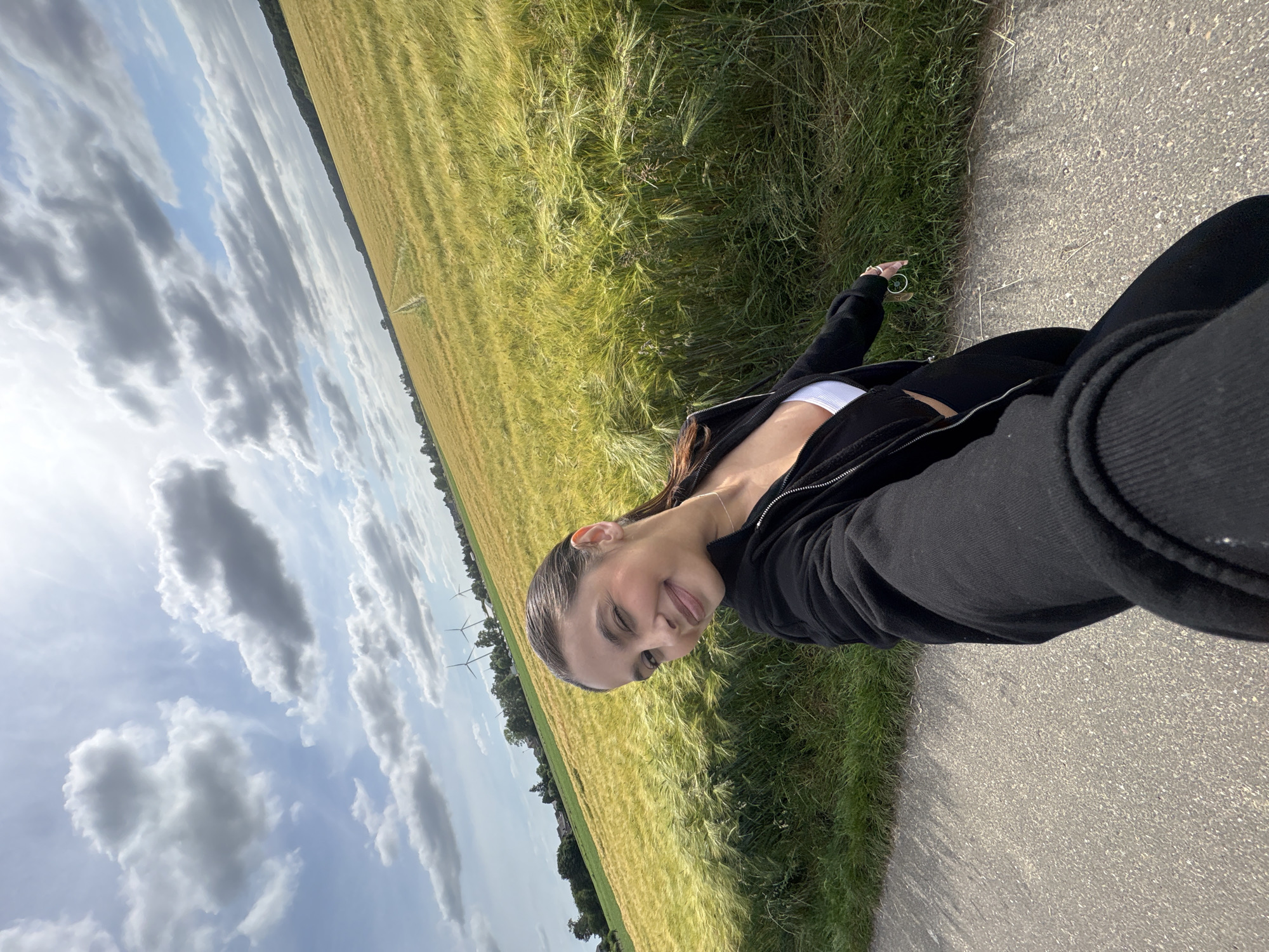 A young adult female is depicted in a selfie, positioned on a paved path with her arm extended towards the camera. She smiles and winks with her left eye, her dark hair pulled back. She wears a black zip-up jacket over a white crop top and black leggings, accessorized with a delicate necklace. To her left and stretching into the midground is an extensive field of golden-green agricultural crops, consistent with barley or wheat, exhibiting gentle undulations and a faint dirt track winding through it. Tall green grass borders the paved path. In the distant background, scattered residential buildings and trees are visible to the left, while three operational wind turbines rise prominently above the horizon to the left of the frame's center. The sky overhead is partly cloudy, displaying patches of bright blue and scattered white and grey cumulus clouds, with strong sunlight illuminating the scene. The overall environment is a rural or semi-rural setting, characteristic of the region of Wegberg, Germany, featuring agricultural fields and renewable energy infrastructure.
