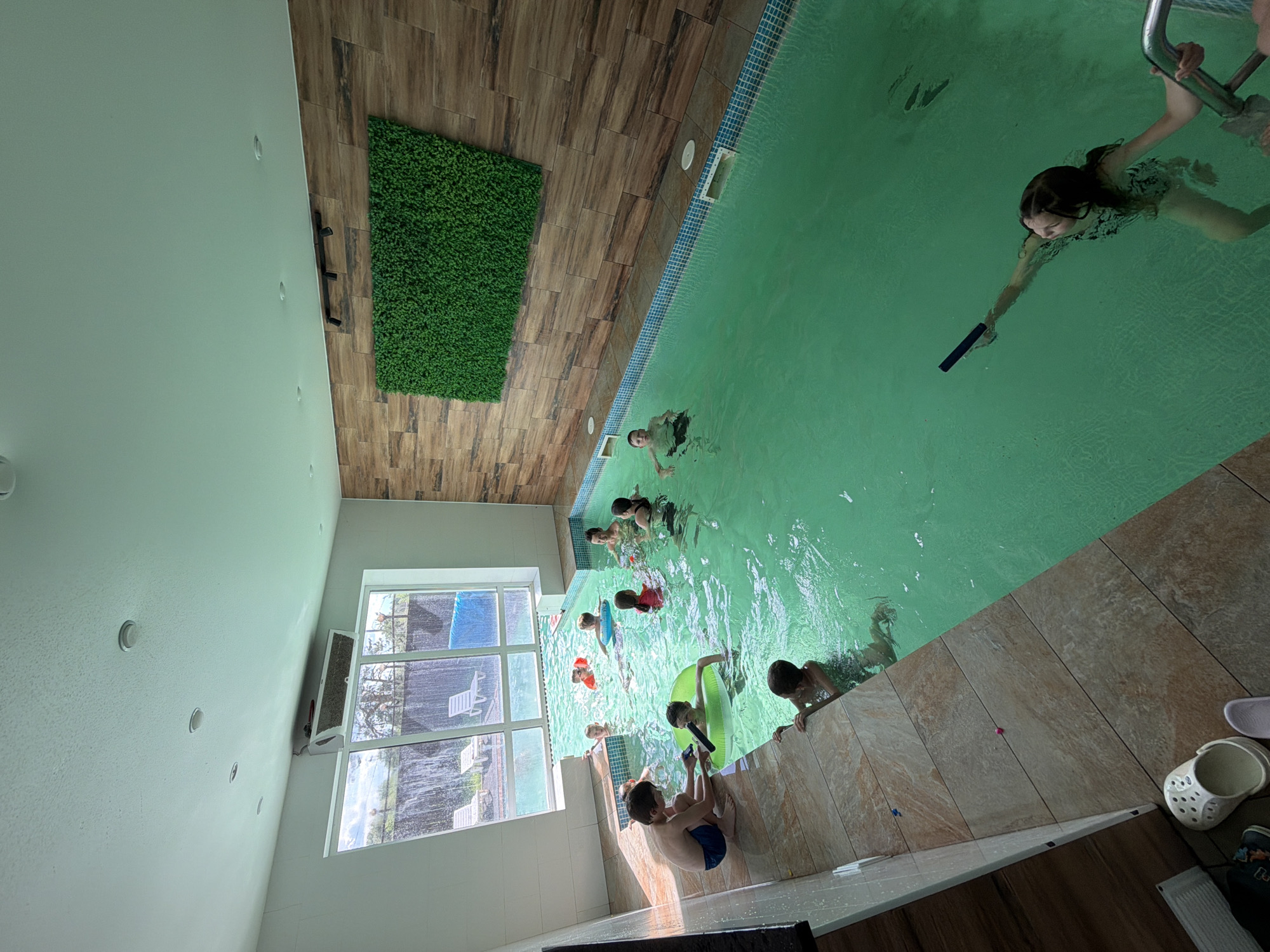 An indoor swimming pool in a facility in Chaikovsky, Russia, is filled with approximately 10 people, mostly children, swimming and interacting. The pool has green-tinted water and is bordered by a tiled edge. One side of the pool area features a large window with lounge chairs visible outside, and a waterfall feature. The walls are decorated with a wood-grain tile pattern and a large artificial green wall. Several children are seen wearing flotation devices, and one child is playing with a long, dark object in the water. The scene appears to be a recreational setting.