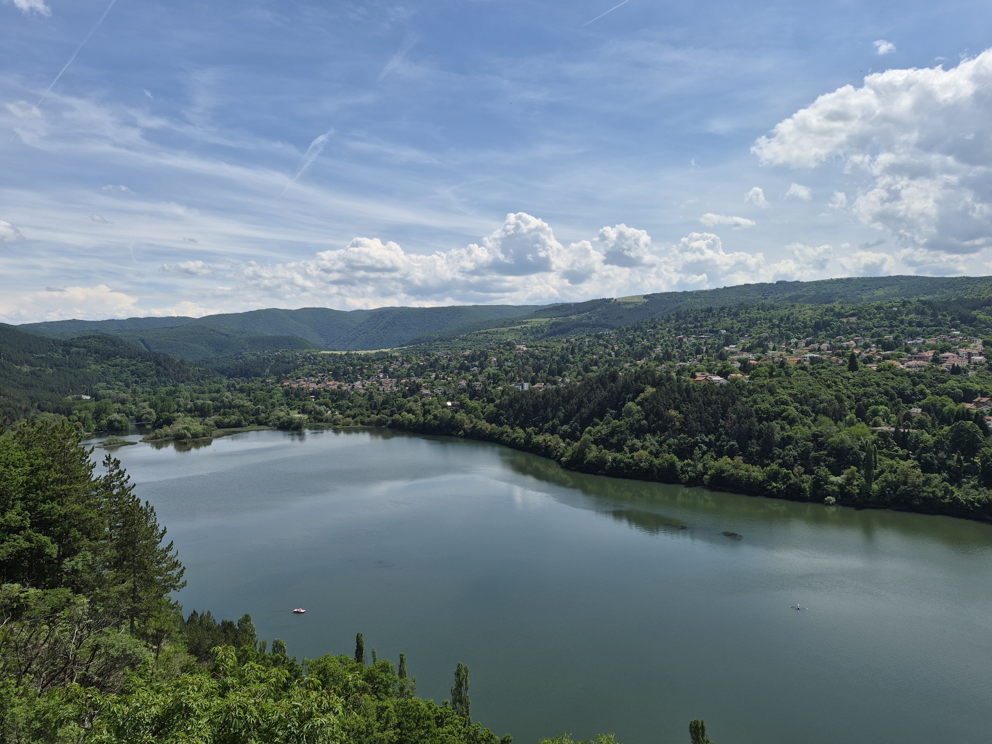 The image captures a panoramic view of a tranquil lake surrounded by lush green hills and a dense forest. A small village or town is nestled among the trees on the distant hills, with houses featuring red-tiled roofs. The sky above is a vibrant blue, dotted with fluffy white clouds and wispy contrails. The lake itself appears calm, reflecting the sky and surrounding greenery. In the foreground, the edge of a steep, tree-covered slope is visible. Two small boats are present on the water: one a pedal boat near the left bank, and another a small rowing boat with a single occupant in the middle of the lake. The overall scene conveys a sense of natural beauty and peaceful coexistence between the settlement and its environment.