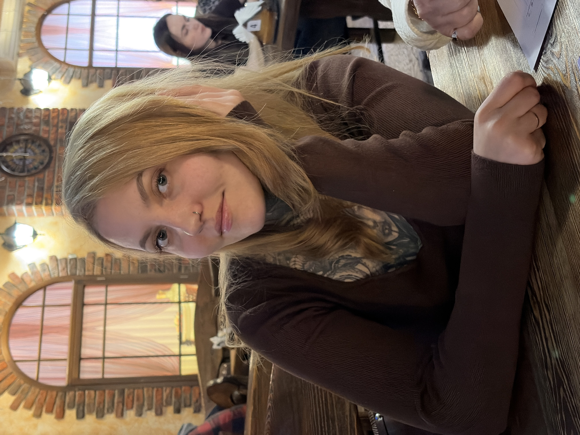 A woman with fair skin, blonde hair, and a nose piercing is seated at a wooden table, likely in a restaurant or cafe. She wears a long-sleeved brown shirt and has tattoos visible on her neck. Her left hand is resting on the table. The background shows two arched windows with brick framing, and what appears to be a clock mounted on a brick wall. Another person is seated at a table in the background. The scene appears to be in a building with distinctive architectural elements, possibly a restaurant or cafe. The location is Chaikovsky, Russia.