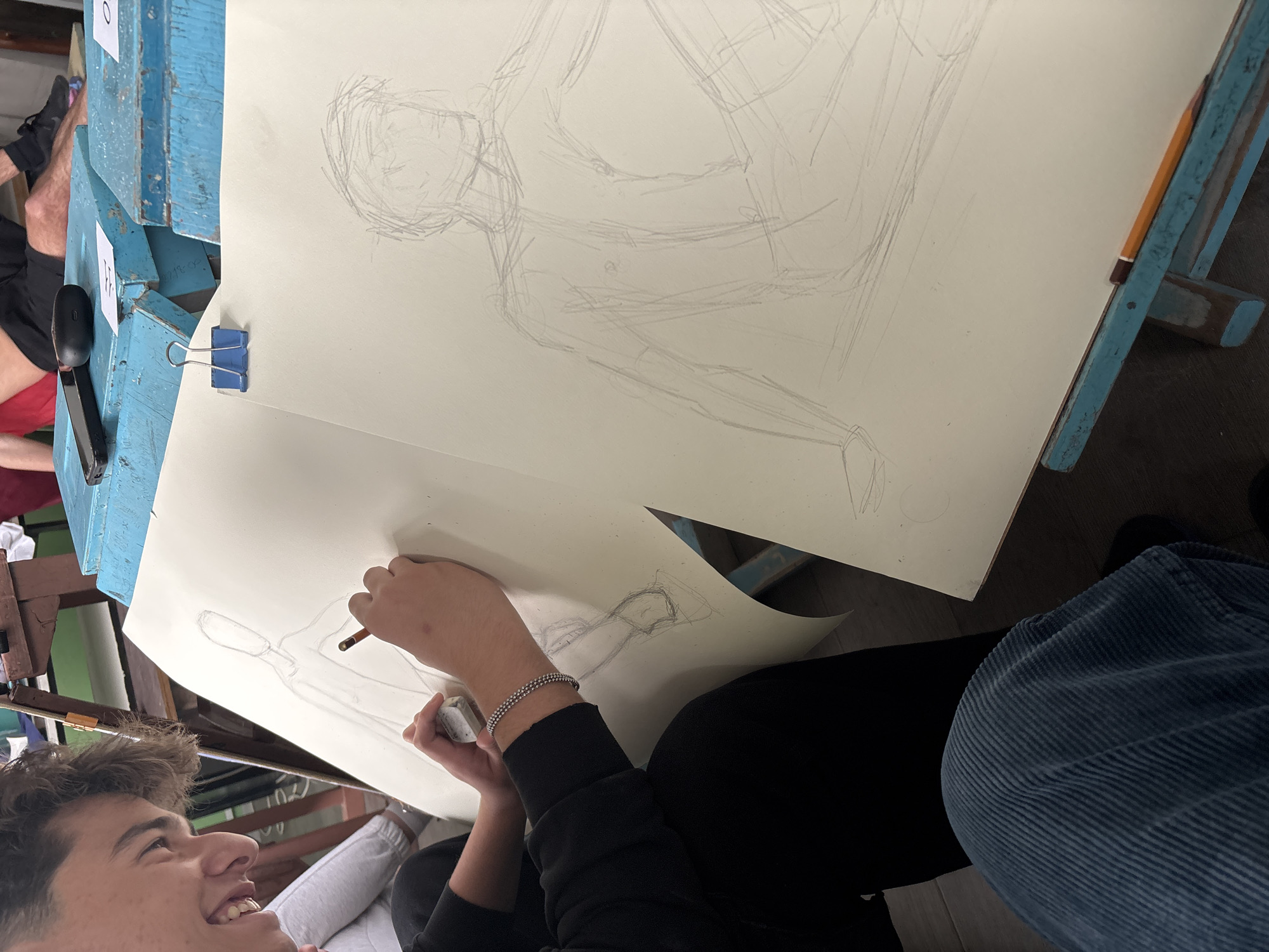 The image depicts an indoor art class or workshop in Troyan, Bulgaria, seen from a high-angle perspective looking down at a student. In the foreground, a young person with short, dark hair and a wide smile is engaged in drawing a human figure on a sheet of paper. They hold a pencil in their right hand and an eraser in their left. A silver, double-strand beaded bracelet is visible on their left wrist. They are wearing a black long-sleeved top, and part of their dark blue corduroy pants is visible in the lower right.
Dominating the center of the frame is a large sheet of white drawing paper, held by a blue binder clip to a light blue wooden drawing board or easel. On this paper, a faint, initial pencil sketch of a human figure in a seated or reclining pose is visible, suggesting a figure study in progress. Another yellow and brown pencil rests on the edge of the blue wooden board to the right. On the blue wooden structure to the left, a black staple gun and two white cards, one marked with "0" and the other with "F", are visible. Part of another person's legs and foot, wearing a black sneaker, can be seen in the background. The overall scene suggests a vibrant and focused art environment.