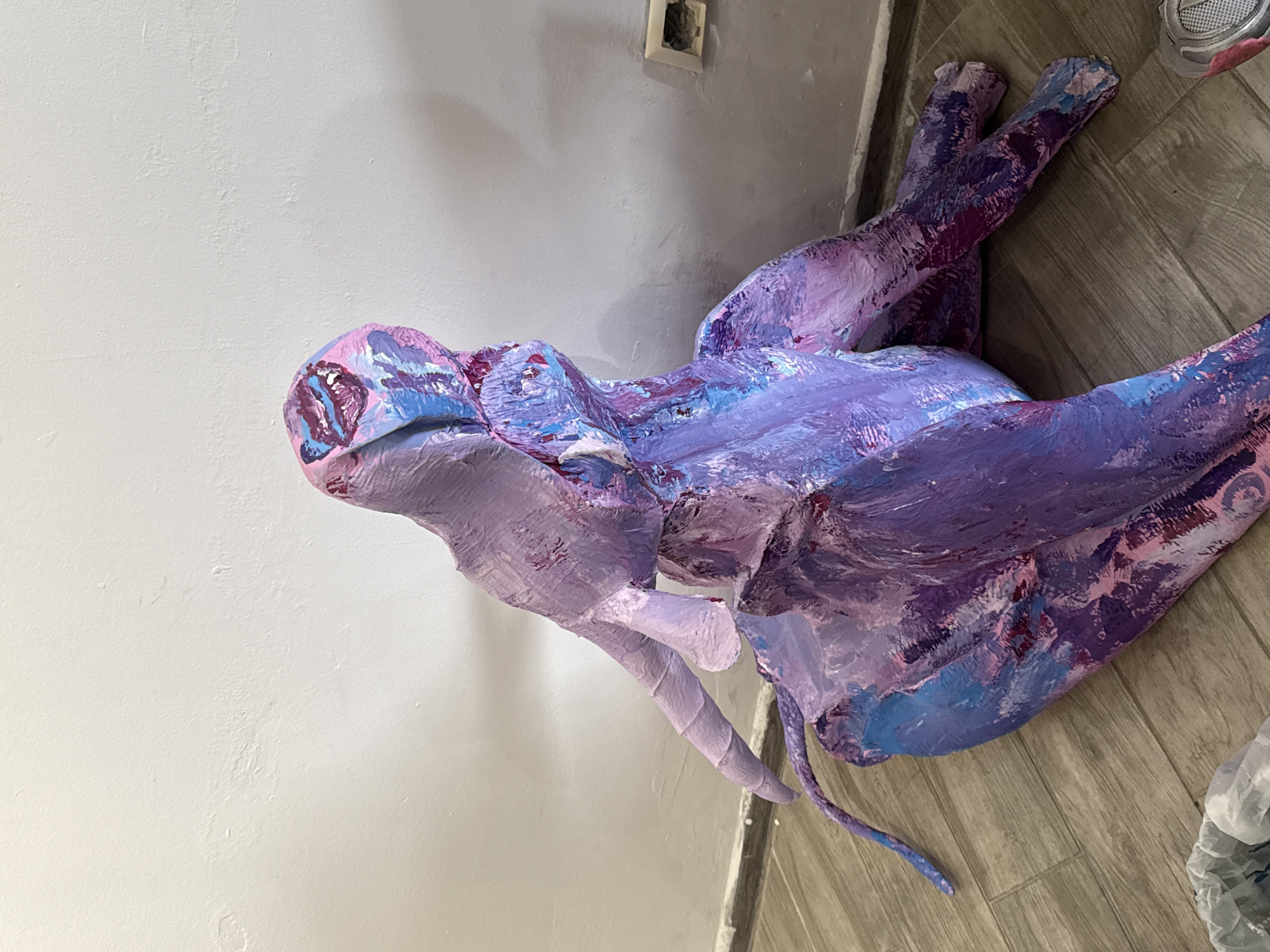 The image shows a sculpture of a dragon-like creature, predominantly painted in shades of purple, pink, and blue. The sculpture has a stylized head with two curved horns and a wide body. It appears to have four limbs and a tail. The texture suggests it is made from a material with a rough surface, possibly paper mache or a similar substance, thickly coated with paint. The sculpture is placed indoors, with a white wall visible to the left and a gray wall with an electrical outlet above. The dragon rests on a light brown, wood-grained floor. The setting does not suggest any connection to Batumi, Georgia. The image contains no people.