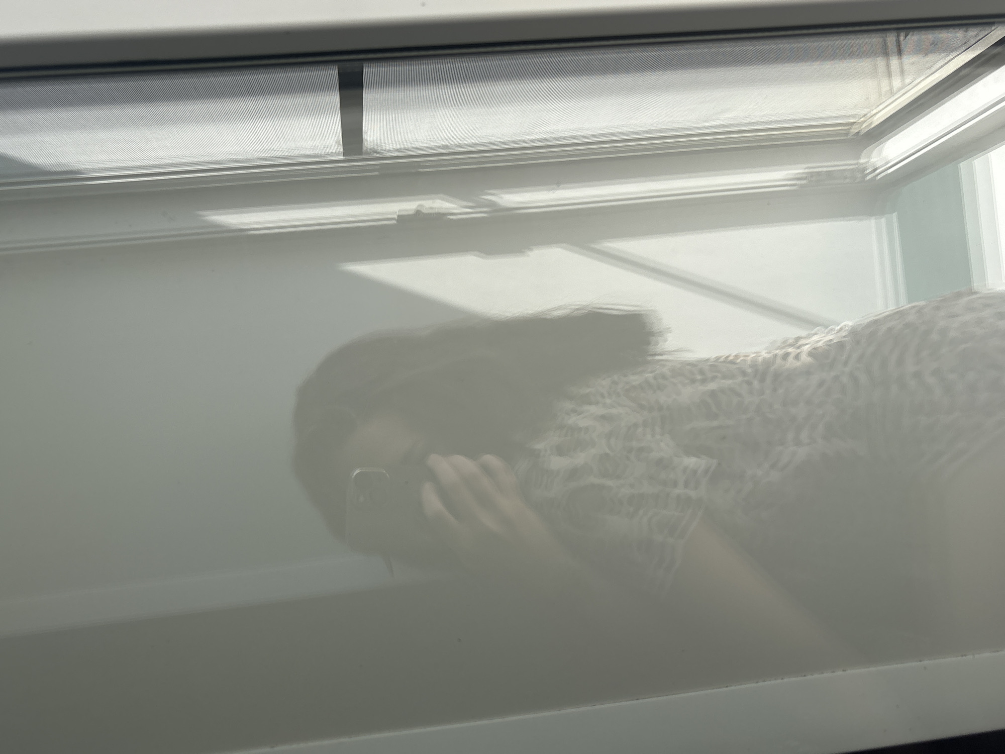 A reflection of a person holding a mobile phone is visible in what appears to be a mirror or a reflective surface, positioned in front of a window with white blinds. The person is wearing a patterned shirt. Sunlight is streaming through the window, casting shadows on the wall and the reflective surface. The scene is indoors, and the setting does not provide any specific indicators to determine its location in the city of Inskoy, Russia.