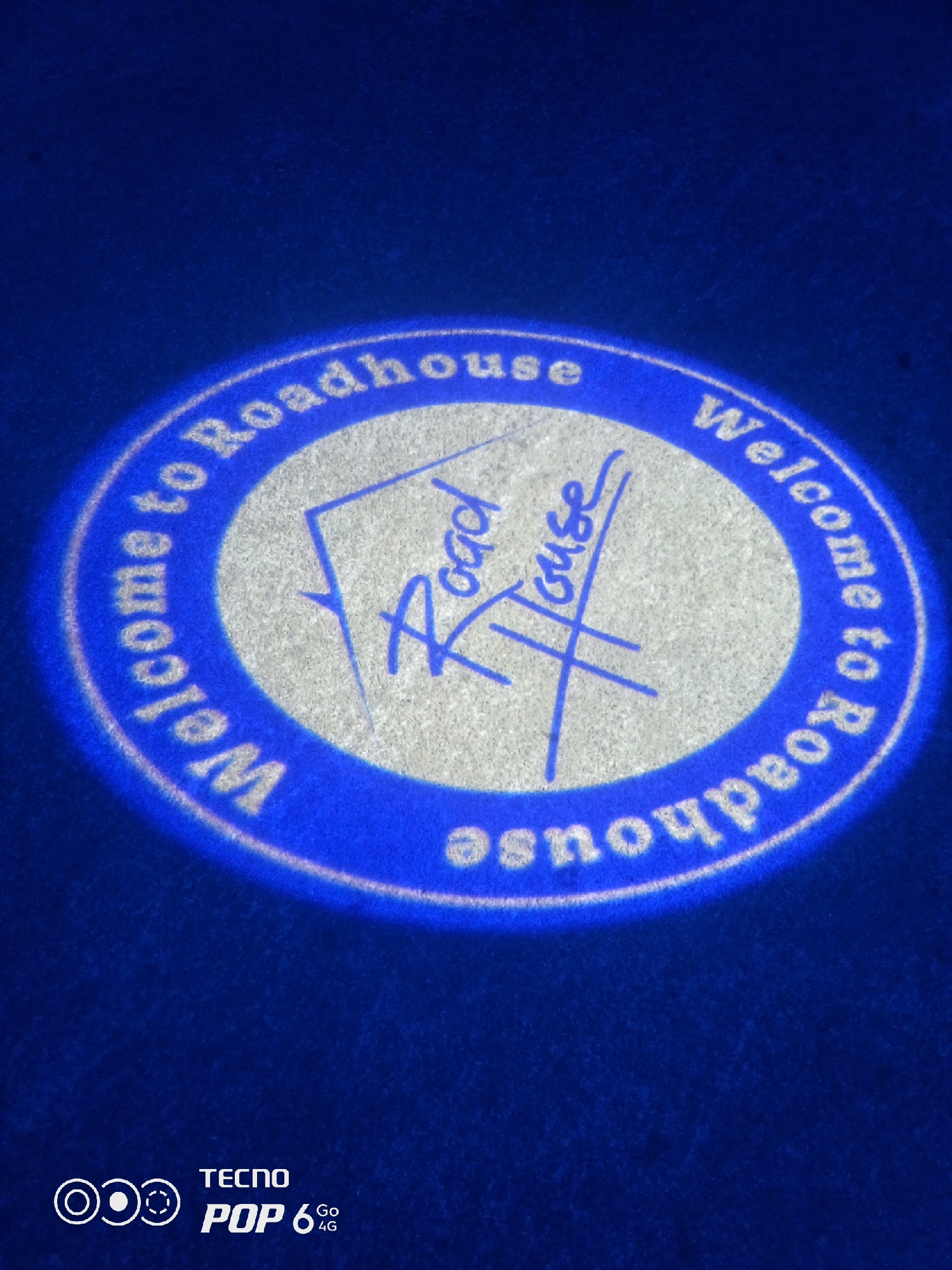 The image displays a close-up, low-angle shot of a projected logo on a textured blue surface, likely a mat or carpet. The projected logo is circular and reads "Welcome to Roadhouse" in white text, with a stylized representation of a house in the center. The projection is slightly blurred and has a halo effect due to the lighting. The bottom right corner of the image features the "TECNO POP 6 Go 4G" logo, indicating the device used for capture. The scene is set in Howayi, Nigeria, though no specific city landmarks are visible.