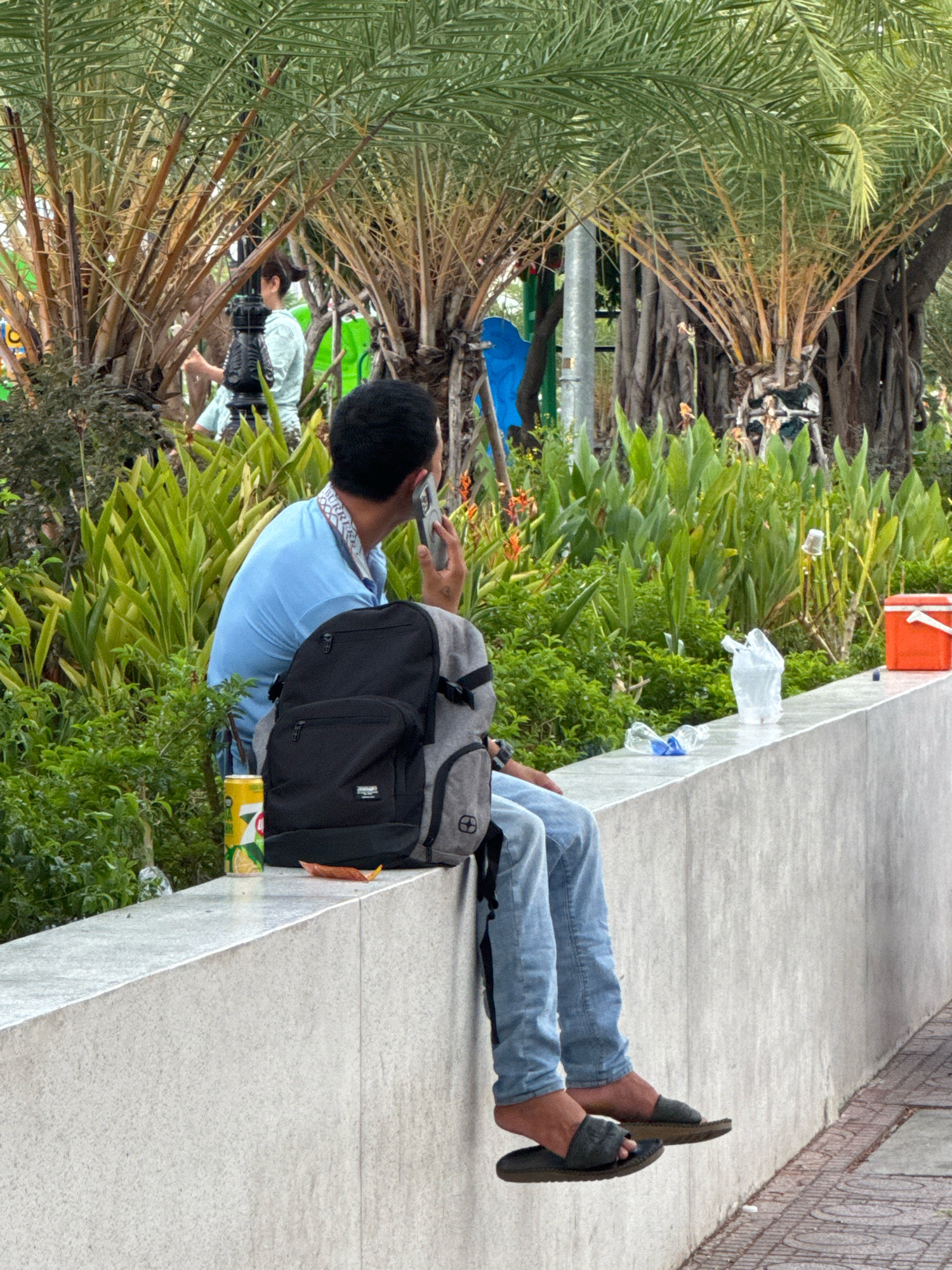 In Nha Trang, Vietnam, a person with dark hair, wearing a light blue collared shirt and light blue jeans, sits on a concrete wall. They are holding a smartphone to their ear and talking, with a grey and black backpack resting on their lap. A yellow can of 7 Up is placed on the wall to their left, and a small, brown leaf is near the backpack. The person is wearing dark colored flip-flops. The background features lush green foliage, tall palm trees with brown trunks, and a blurry image of another person in the distance. To the right of the main subject, a white plastic bag is on the wall, and further to the right, an orange cooler is visible. The wall itself is a light grey concrete structure.